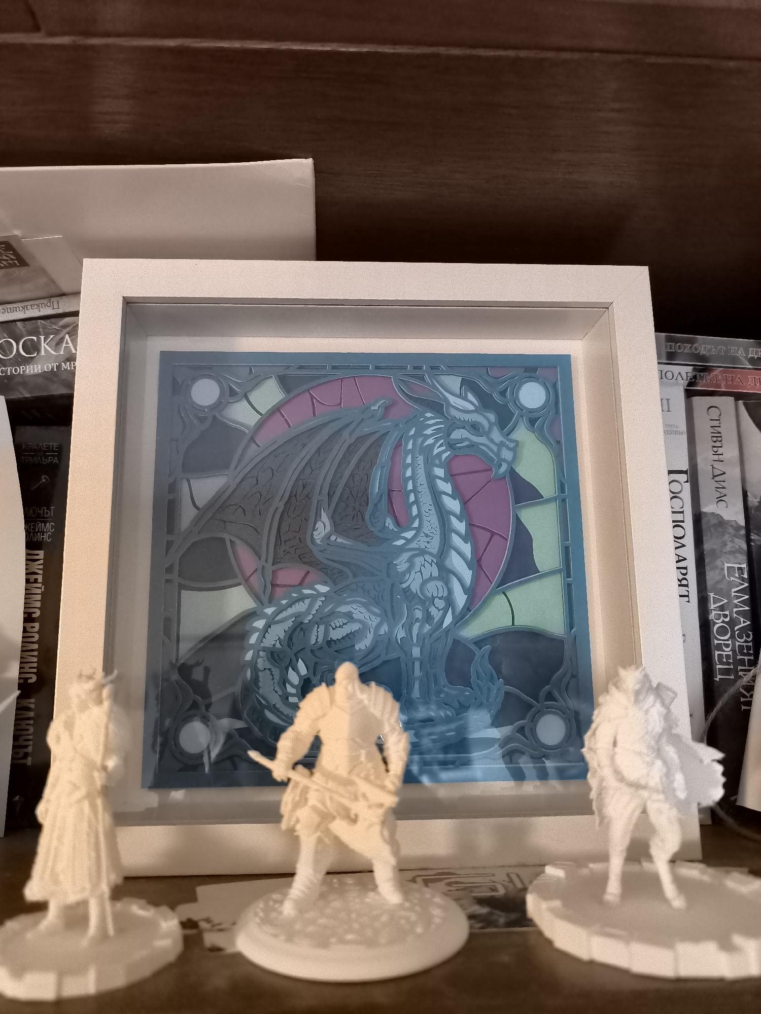 A close-up shot shows a framed piece of artwork depicting a dragon in a stained-glass style, positioned on a shelf. The artwork is square, with a white frame. The dragon design features shades of blue, grey, pink, and green. In front of the framed artwork, three white miniature figures are arranged on the shelf. Behind the framed artwork are several books, oriented vertically, with visible titles in what appears to be Cyrillic script. The shelf itself is brown and appears to be made of wood. The environment is indoor and seems to be a bookshelf. The image does not show any location in Burgas, Bulgaria.