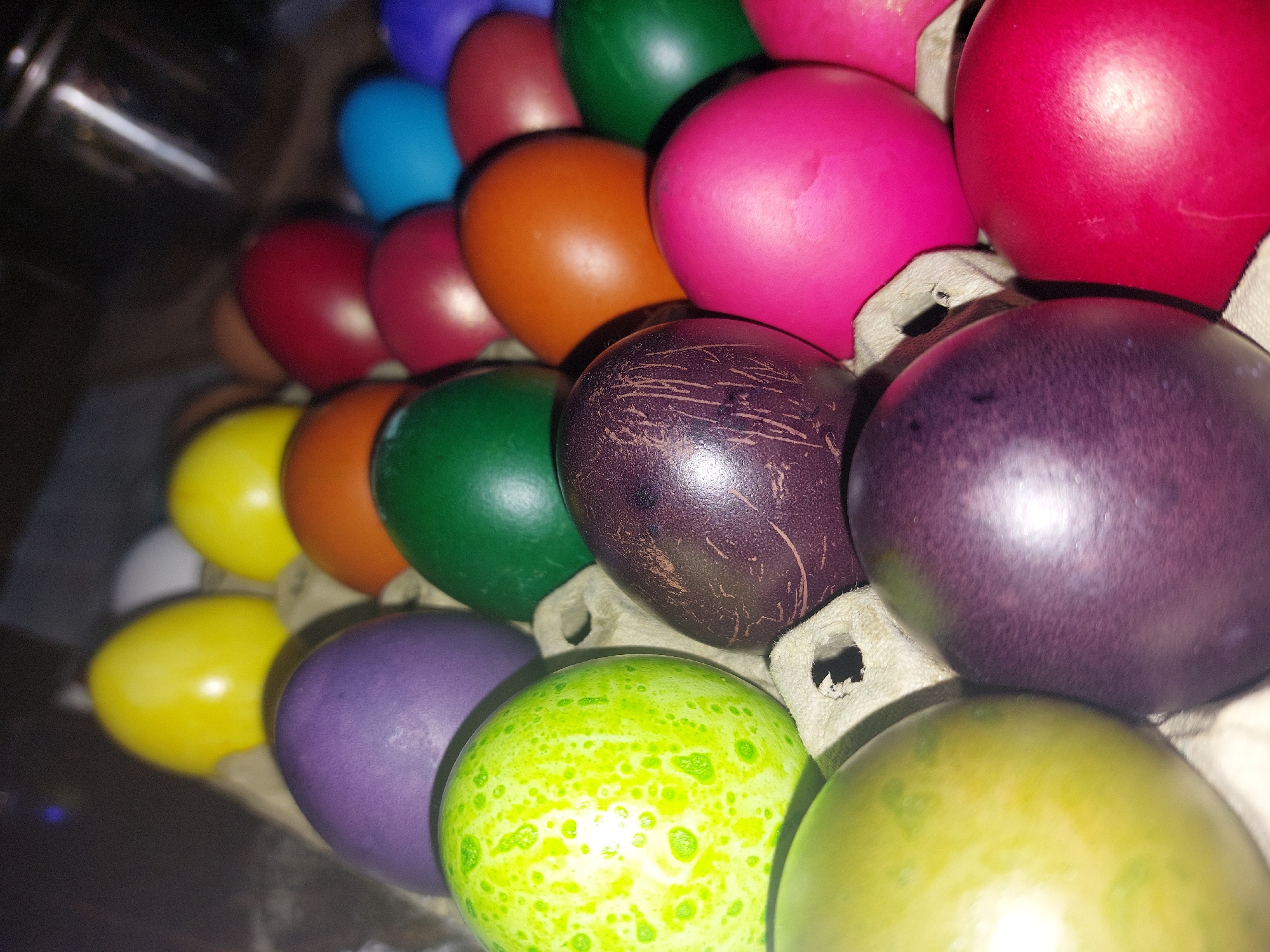 A close-up shot depicts a collection of brightly colored Easter eggs arranged in an egg carton. The colors include red, pink, orange, yellow, green, blue, and purple. Some eggs have speckled patterns. The lighting casts a glossy sheen on the eggs, highlighting their smooth surfaces. The background is blurry and indistinct.