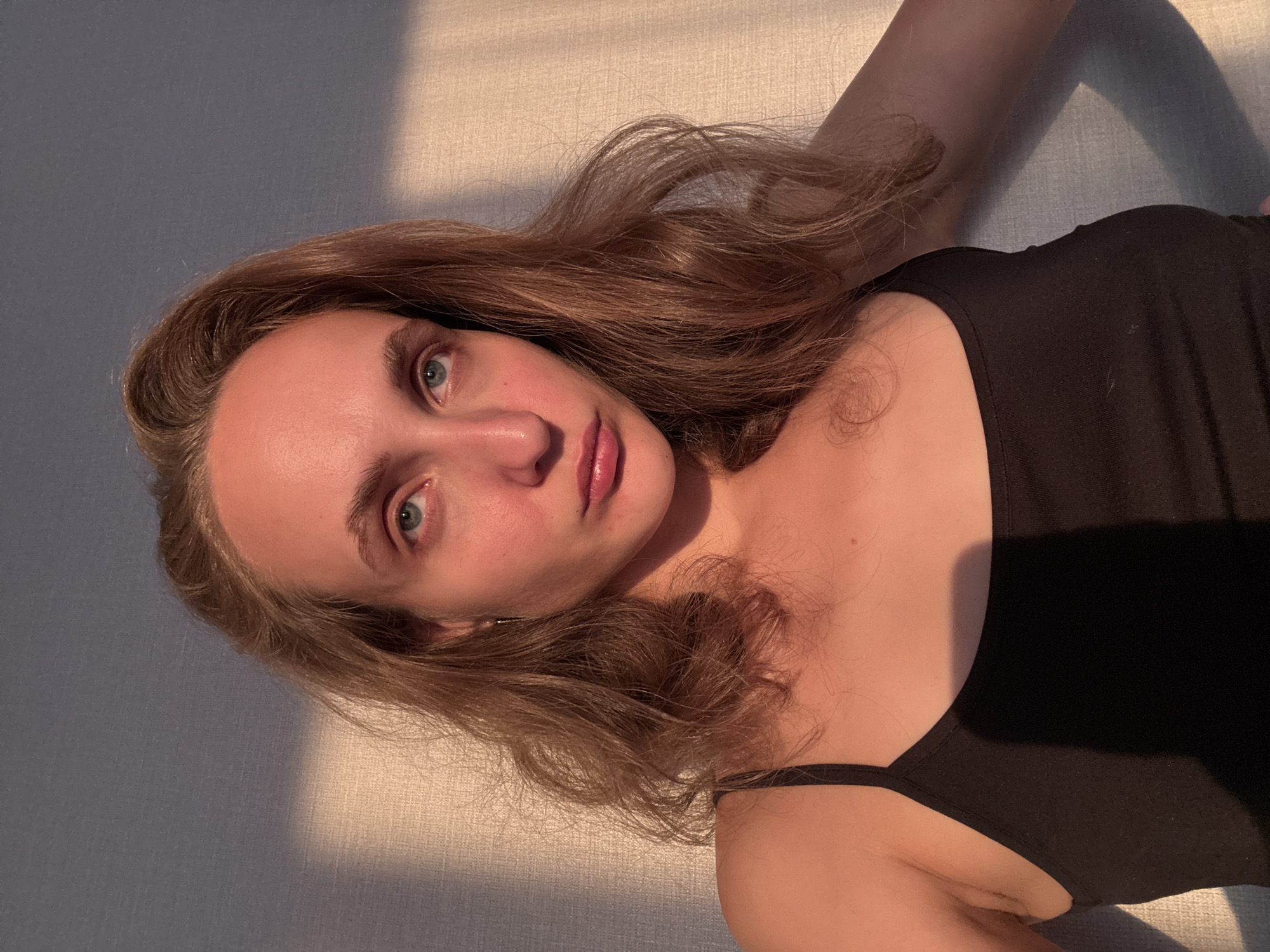 A young Caucasian woman with light brown, wavy hair is shown from the chest up, facing the camera. She is wearing a black tank top. Her skin is flushed, possibly from the sun or a warm environment. Her facial expression is neutral, with her lips slightly pursed. The background consists of a textured wall with natural light casting shadows across it. The image appears to have been taken indoors, possibly in a home, as indicated by the lighting and background. There are no identifiable landmarks or city-specific features visible in the image.