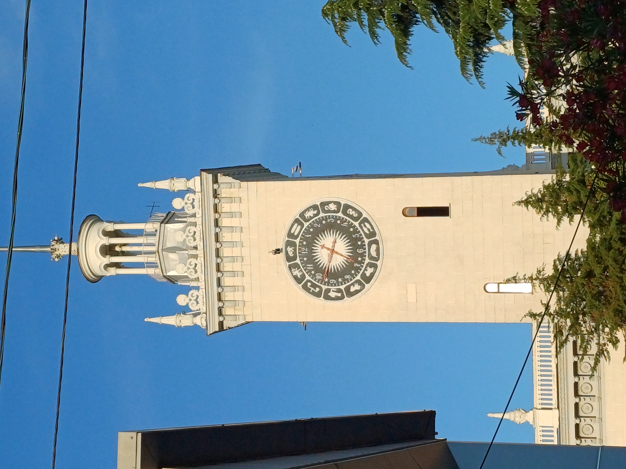 The image shows a close-up of the Rotunda at the Primorsky Boulevard in Sochi, Russia. The Rotunda is a prominent architectural landmark known for its distinctive clock face, which displays zodiac signs around its perimeter. The clock's hands are visible, suggesting a specific time. The sky is a clear, vibrant blue, indicating fair weather, likely during the daytime. Green foliage from nearby trees frames the top right of the image, with some red flowers adding a splash of color. The building itself is light-colored, possibly stone or stucco, with intricate white decorative elements, especially on the spire and the building's facade. Several dark wires are visible in the foreground, running vertically, possibly power lines or telecommunication cables. In the lower part of the image, a dark, flat roof of another structure is visible, providing a contrasting element to the sky and the Rotunda. The overall impression is a bright, sunny day in a picturesque location, highlighting a significant architectural feature of Sochi. There is no visible text in the image.