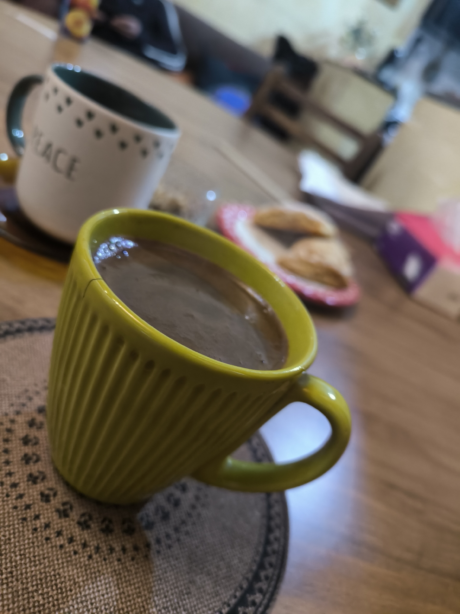 A close-up shot of a breakfast scene on a wooden table. A bright yellow ribbed mug filled with coffee sits on a woven mat in the foreground. Slightly behind, a white mug with the word "PEACE" written on it is partially visible. A small plate with pastries is placed behind the mugs. A box of tissues is further back, partially out of focus. The environment suggests a home setting. There are no people visible in the frame. The location in the city of Tbilisi, Georgia cannot be determined.