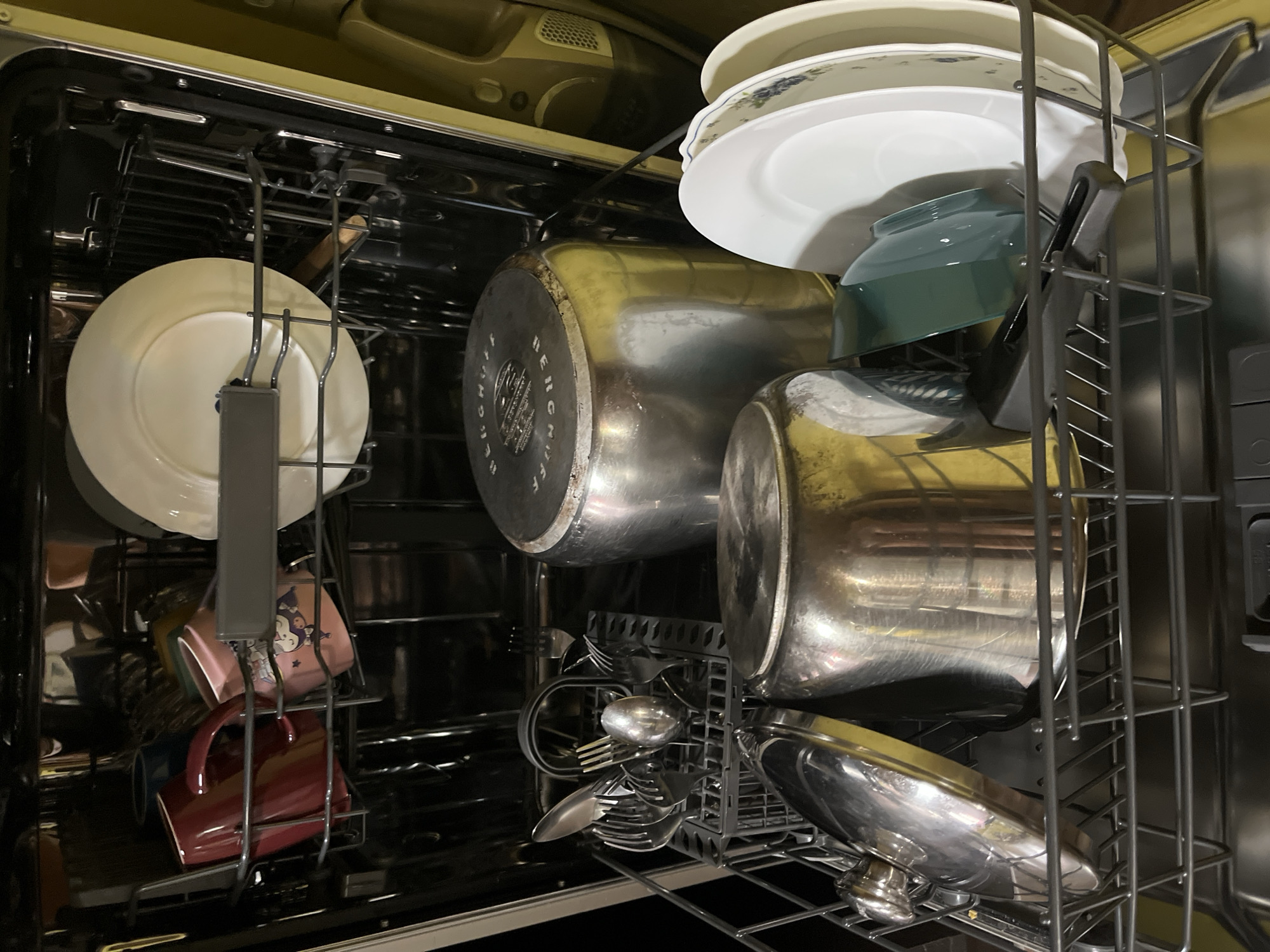 The image shows the interior of a dishwasher filled with dirty dishes. On the top rack, there are several mugs, including a red one and a pink one with a cartoon character, along with a white plate. On the bottom rack, there is a silverware basket containing forks, spoons, and knives. Also on the bottom rack are two large metal pots, one with a lid, and several white plates. A green glass bowl is also visible. The dishwasher appears to be a standard model with wire racks. The lighting is dim, suggesting the photo was taken inside a dimly lit room or at night. There is no information in the image that would suggest a specific location in Tarnivtsi, Ukraine, or any other city. The scene depicts everyday household items in a domestic setting.