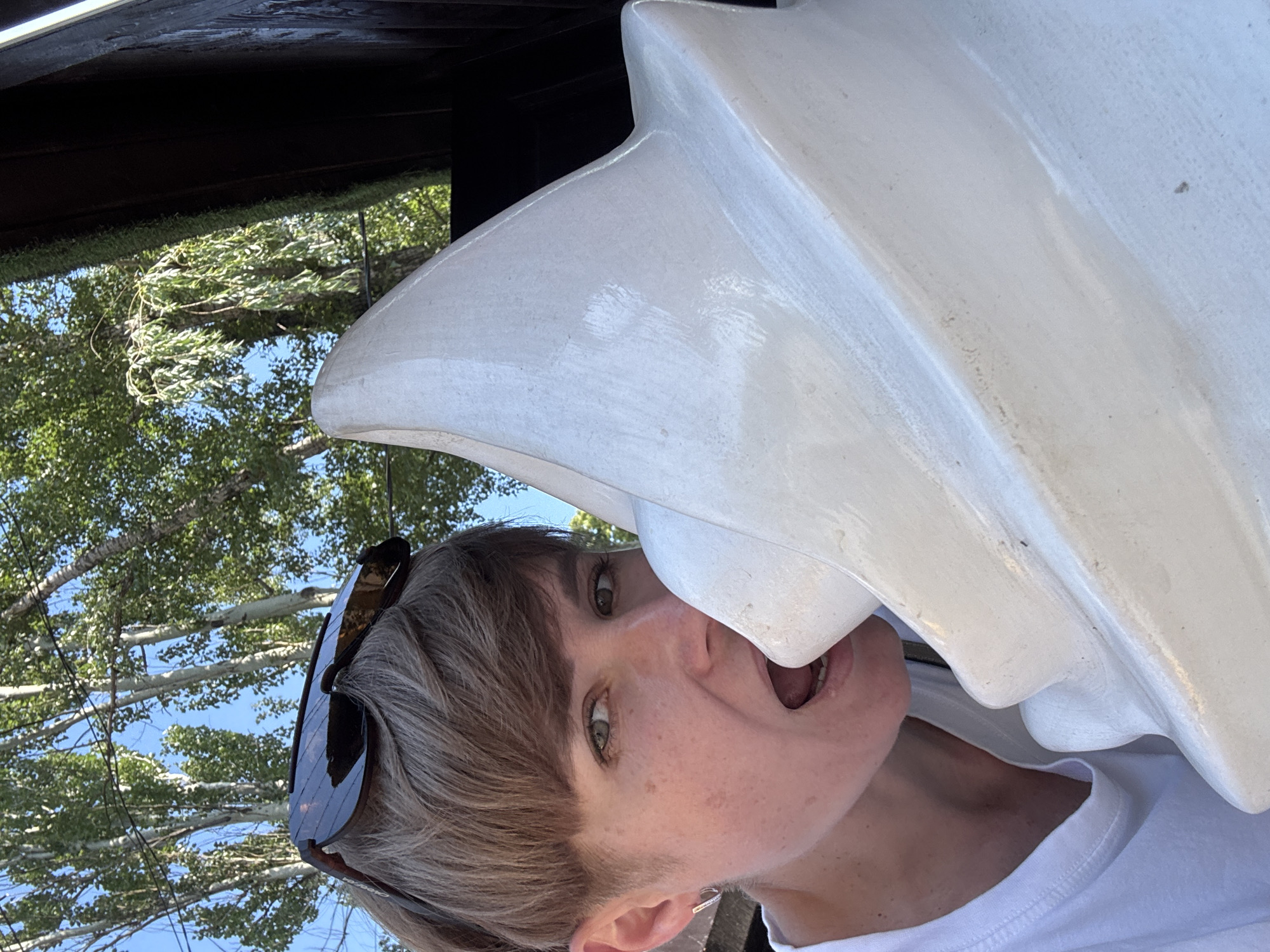 An unidentified individual is pictured in an outdoor, possibly urban, setting, with trees and a bright blue sky visible in the background. The individual, who appears to have short, brown hair and is wearing a white t-shirt and sunglasses, is positioned in the foreground, leaning in towards a large, white, soft-serve ice cream cone sculpture. The person's mouth is open, and they are licking the sculpture. The overall lighting suggests it is daytime, with the sun casting shadows and highlighting the textures of the sculpture. The scene is captured in a close-up perspective, focusing on the interaction between the individual and the ice cream cone.