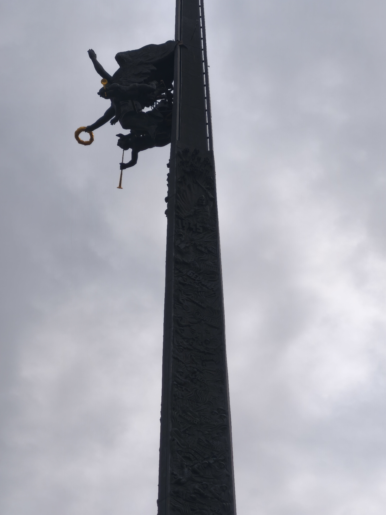 The photograph shows a low-angle, vertical shot of the Victory Monument in Moscow, Russia, on a cloudy day. The monument is a tall, dark obelisk with detailed carvings along its base. At the top of the monument, silhouetted against the sky, are bronze sculptures of figures. One figure, reaching upwards, is holding a golden laurel wreath. Another figure below is blowing a golden trumpet. The overcast sky creates a muted, grayish backdrop for the dark monument and its sculptures.
