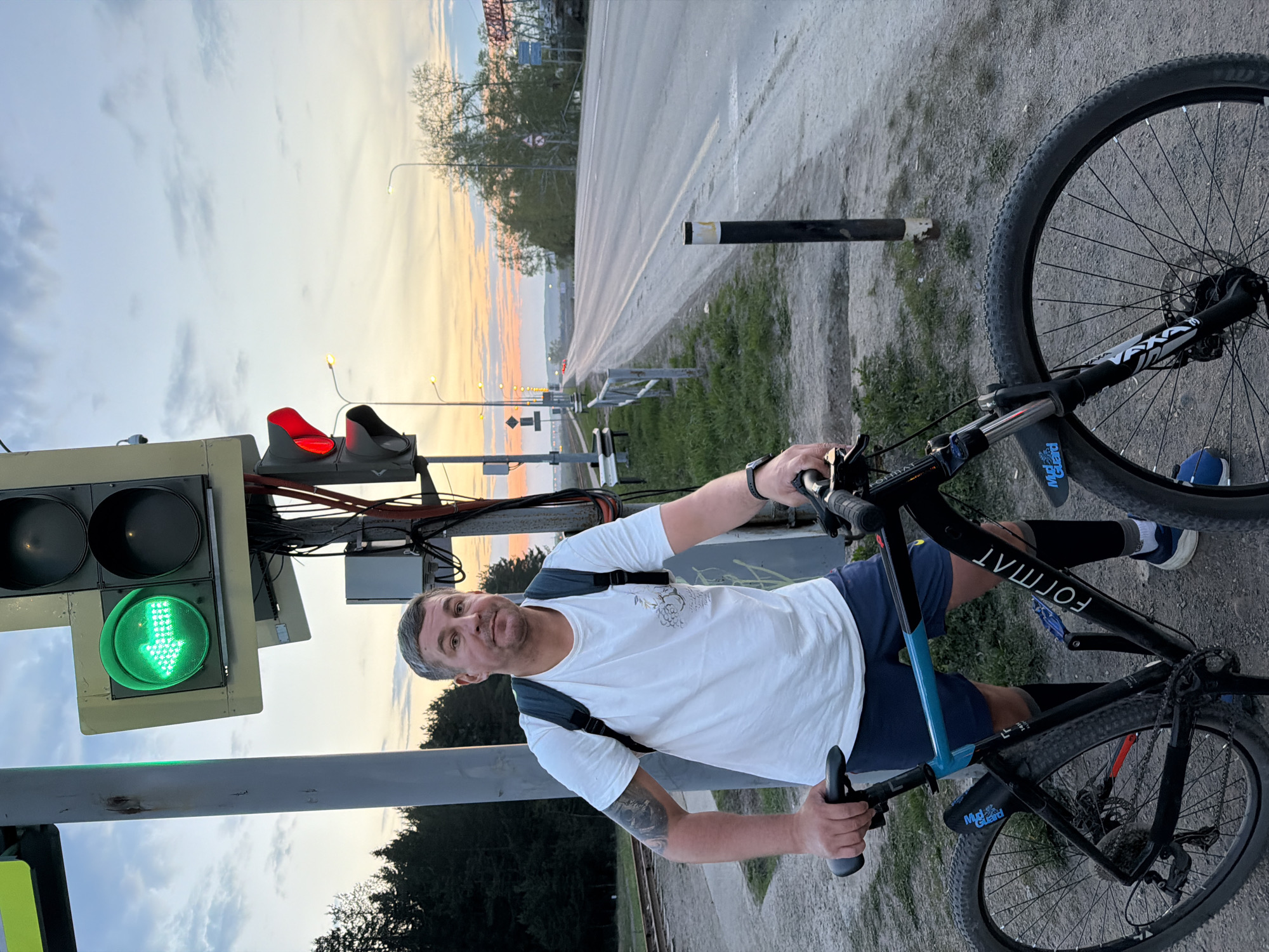 A man with short gray hair, wearing a white t-shirt and blue shorts, stands with a black and blue bicycle next to a traffic light. The traffic light shows a green arrow indicating a left turn. A red traffic light is visible above the green arrow. The man is looking at the camera and holding the handlebars of the bicycle. Behind him is a road and the sky is illuminated by the colors of the sunset. The photograph was taken in Chaikovsky, Russia.