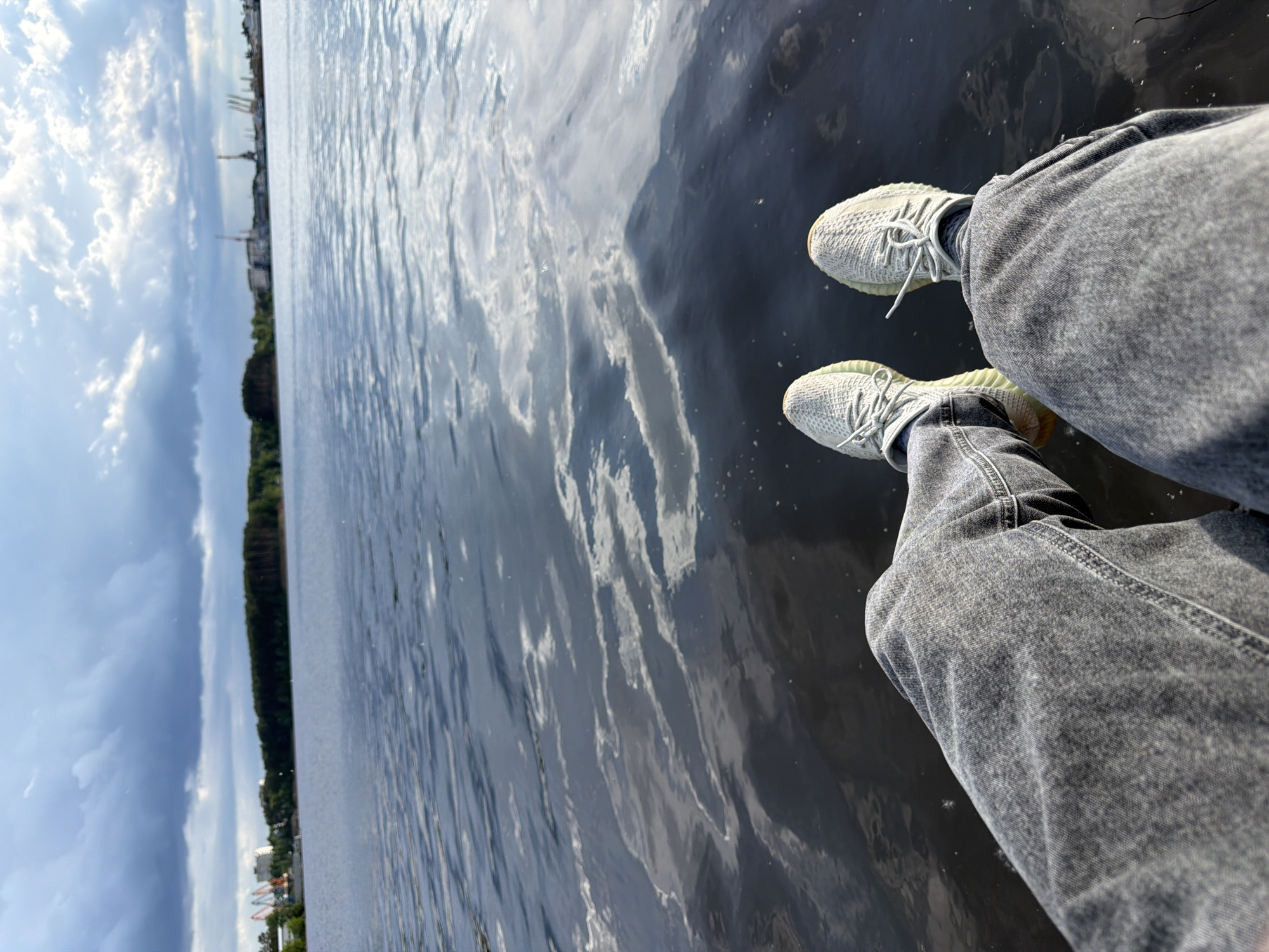 A low-angle, rotated perspective shows a person sitting on a structure over water. The person is wearing grey jeans and light grey sneakers with white laces, with their legs hanging over the edge into the water. The water reflects the sky, which is partly cloudy with varying shades of blue and grey. In the distance, there is a treeline and beyond that, what appears to be a cityscape with buildings and structures. Based on the appearance of the city and the request that specifies Chaikovsky, Russia, it can be inferred that the scene is located in Chaikovsky, Russia, or a nearby area. There is no obvious movement or interaction occurring in the scene.
