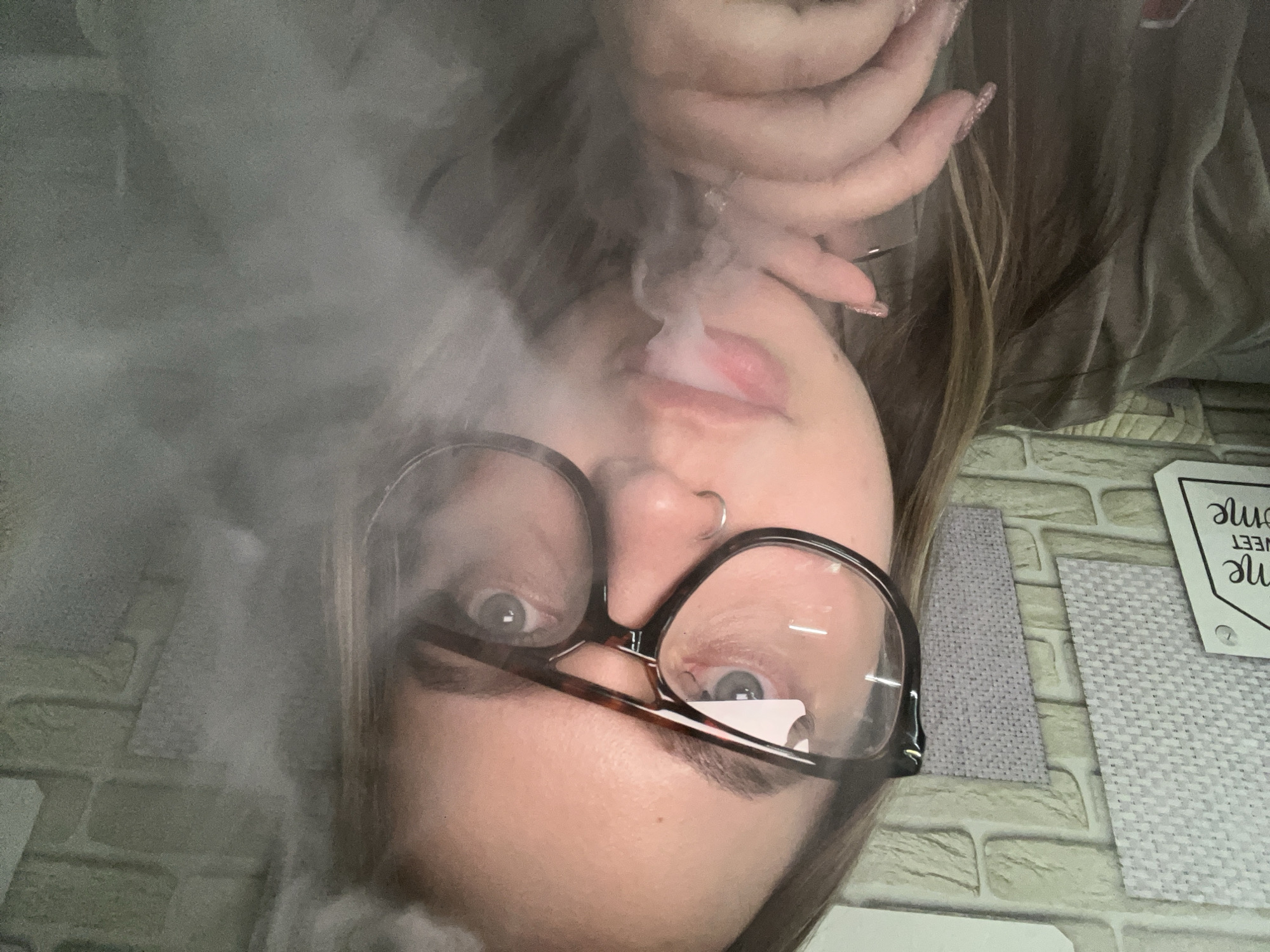 An inverted, close-up indoor portrait taken in Port-Katon, Russia, features a person exhaling a dense cloud of white vapor or smoke. The individual's face, oriented upside down, is largely visible through the swirling smoke. They wear dark-rimmed glasses, which reflect some light, and have a small silver hoop nose piercing. Their eyes are open, looking directly forward. Pinkish lips are slightly parted as the vapor exits. One hand, adorned with long, possibly glitter-tipped fingernails, is visible near the person's mouth and hair. The hair is long and appears to be a mix of brown and lighter streaks.
The background consists of a textured surface, resembling light grey or beige brickwork wallpaper or tiles. On this surface, to the lower right, are two rectangular fabric swatches or coasters in varying shades of grey. A white, house-shaped tag or sign is also visible, bearing the partial text "...SWEET HOME". The overall lighting is dim, emphasizing the contrast of the bright vapor against the darker tones of the subject and background. The focus is sharp on the person's face and the immediate vicinity of the smoke.