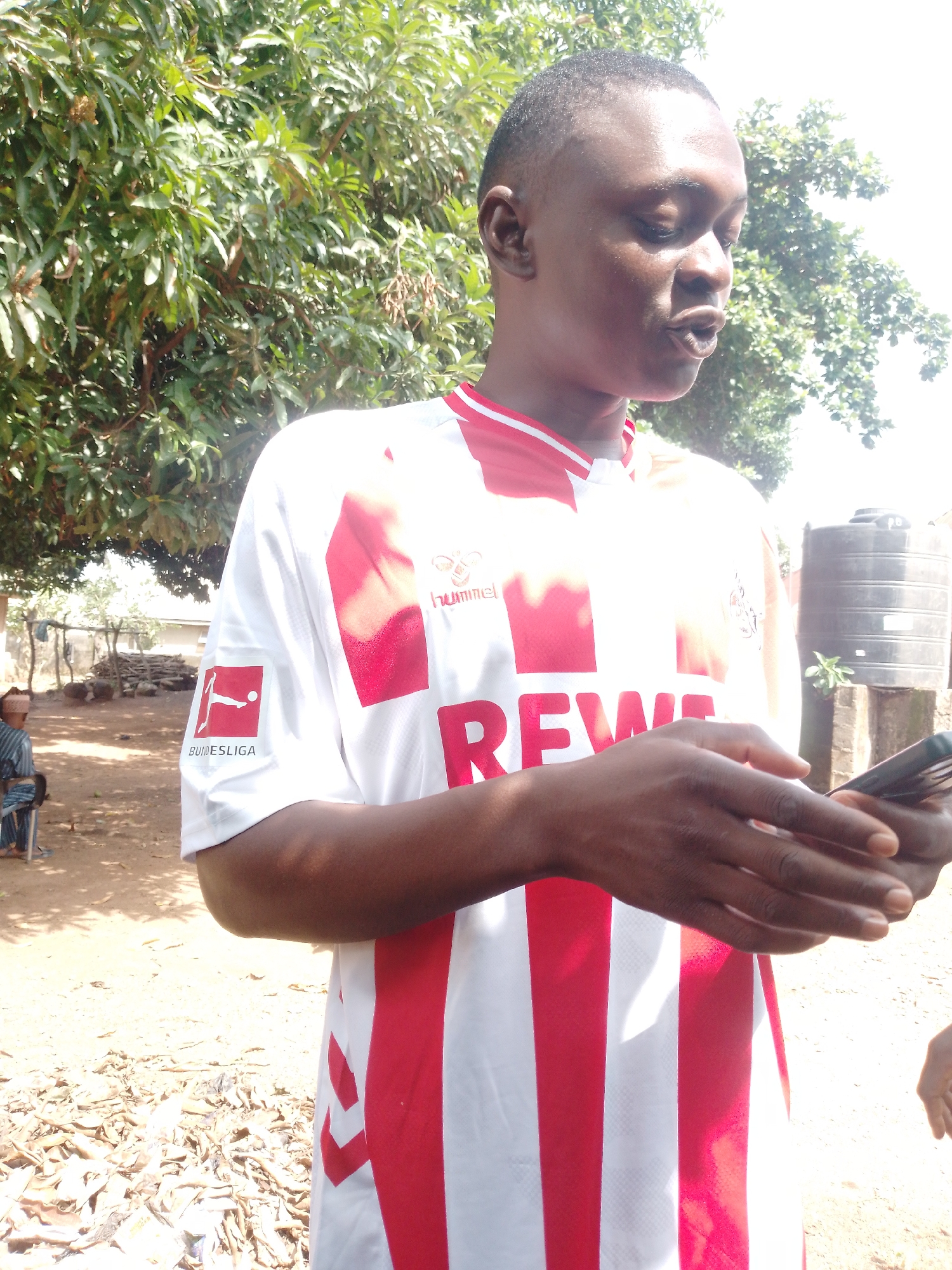 A young Black man wearing a white and red striped soccer jersey with a Bundesliga logo on the left chest and the word "REWE" in red letters across the front stands in front of a blurred background of trees and a black water tank. He is looking down and to his right, with his mouth slightly open as if speaking. His hands are visible, holding a dark-colored mobile phone. The ground is covered in dirt and dry leaves. In the background, a person is seated on a chair to the left, and there are some wooden structures and debris. The lighting is bright, suggesting it is daytime. The location is listed as Unknown, Unknown.