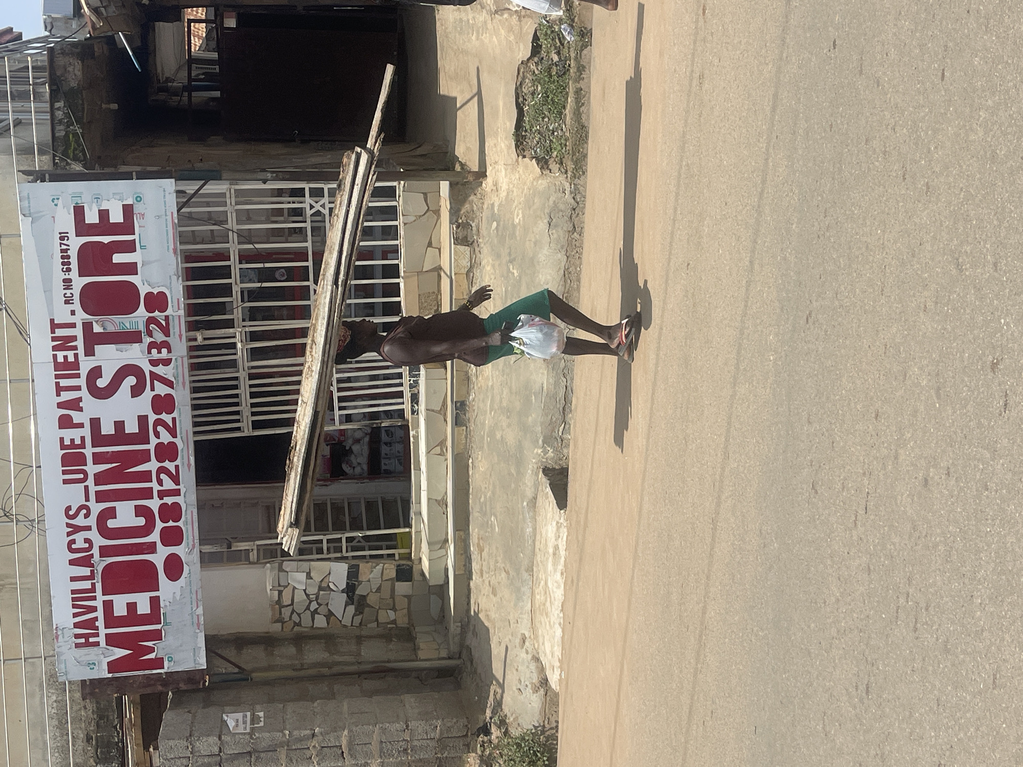 A man is walking on a street in Abuja, Nigeria, carrying a load of wooden planks on his head. He is wearing a brown tank top and green shorts, and he is carrying a white plastic bag in his left hand. The man is walking in front of a store with a sign that reads "HAVILLACYS_UDE PATIENT MEDICINE STORE". The store appears to be a local pharmacy or general store. The street is paved, and there are some plants and debris on the side of the road. The overall scene appears to be a typical street view in a Nigerian city.