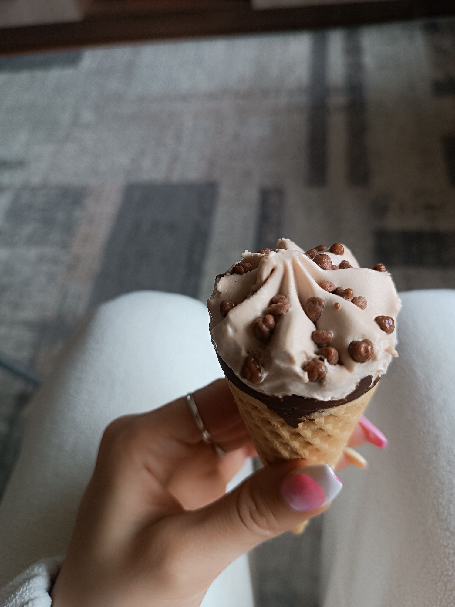 This vertical close-up photograph captures a person's hand holding a single ice cream cone indoors. The hand, with a light to medium skin tone, is positioned in the lower-middle of the frame, gently grasping the waffle cone. The cone is golden-brown and features a dark chocolate lining around its top rim. The ice cream itself is a soft, swirled mound of light brown or beige-colored frozen dessert, likely a chocolate, coffee, or caramel flavor. It is generously adorned with numerous small, round, dark brown crunchy toppings, resembling chocolate crisps or malted milk balls.
The fingers of the hand display manicured nails, which have a distinctive design: a white tip often associated with a French manicure, combined with a vibrant pink color forming a crescent or diagonal shape on the nail bed. A slender, silver-colored ring is visible on the ring finger.
The background is softly blurred, indicating a shallow depth of field, which draws the viewer's attention to the ice cream. Behind the hand, a swath of light-colored fabric, possibly white pants or a blanket, is visible. Further in the background, a patterned carpet or rug with shades of grey and white can be discerned, suggesting an indoor setting. The lighting appears soft and diffused, characteristic of natural daylight. There is no visible text in the image. The scene conveys a relaxed moment of enjoying a sweet treat.