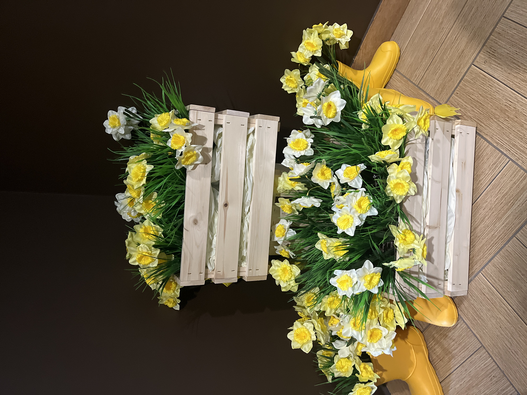 Two wooden crates filled with daffodils and grassy foliage sit on a surface that transitions from dark brown to a light wood-grain pattern. A pair of yellow rubber boots are situated next to one of the crates. The image appears to be taken indoors, as suggested by the artificial lighting and absence of natural elements beyond the plants. The photograph's orientation is tilted, with the crates and background appearing at an angle relative to the horizontal plane. The location is stated as Chaikovsky, Russia.