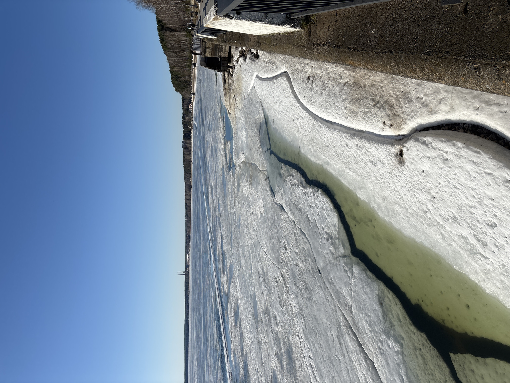 The image depicts a frozen body of water, likely a river or lake, with ice cover displaying cracks and fractures. Some open water or slushy areas are visible along the cracks and near the shoreline. The shoreline consists of a mixture of snow, ice, and exposed earth. In the distance, a treeline is visible, indicating a forest or wooded area along the water's edge. A structure, possibly a bridge or dam, is partially visible in the upper right corner of the frame. The sky is clear and blue.
Given the prompt, this likely depicts the Kama River or Votkinsk Reservoir in Chaikovsky, Russia, during the late winter or early spring thaw.