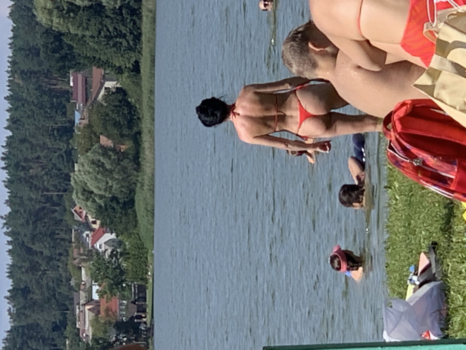 The image displays an outdoor scene at a body of water in Mykhailivka-Rubezhivka, Ukraine, under a clear, sunny sky. The foreground shows a grassy bank with a red backpack and a light-colored tote bag.
In the midground, a minimum of seven individuals are present. A woman with dark hair in a bun stands in the water, facing away from the viewer, wearing a red thong bikini. To her left, one individual is partially submerged wearing a pink visor, and another person floats on their stomach. To the woman's right, the back and shoulders of an individual with light hair are visible, sitting in shallow water, partially obscured by another person's arm. Further out in the water, two additional individuals are partially visible, with only their heads and shoulders above the surface.
The background consists of a sizable body of water. Beyond the water, a settlement is visible, comprising multiple houses with varying roof colors (red, brown, grey) interspersed with trees. A dense coniferous forest forms the horizon behind the houses.
