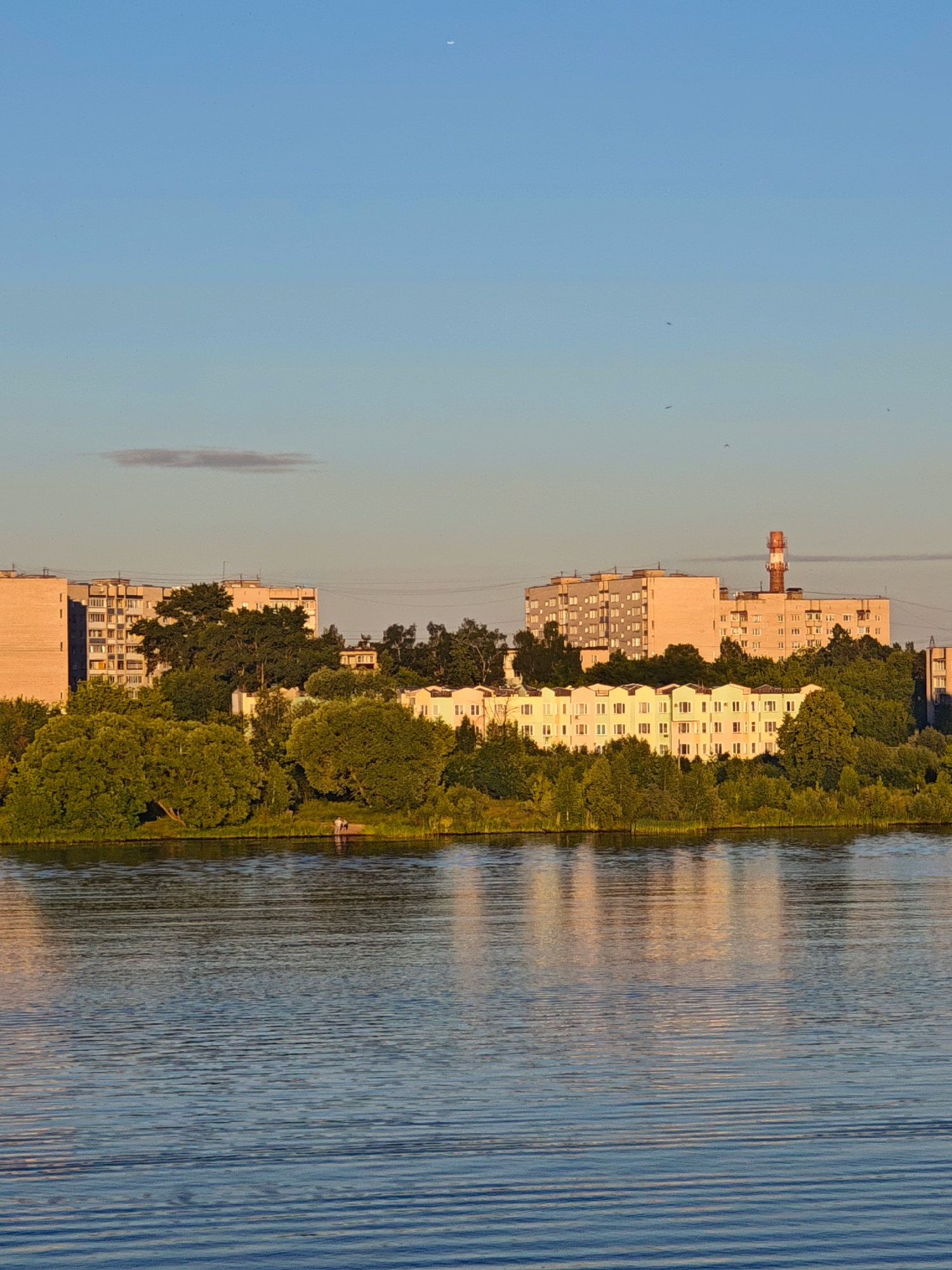 The image captures a serene waterfront scene in Kimry, Russia, likely during the golden hour. A wide body of water, characterized by gentle ripples, occupies the foreground. The water reflects the warm hues of the sky and the buildings. Beyond the water, a lush green treeline runs horizontally across the middle ground, punctuated by several multi-story apartment buildings and a prominent industrial smokestack. The buildings are a mix of styles and colors, with pastel shades dominating some structures and the smokestack exhibiting a red and white striped pattern. The sky above is a clear, soft blue, transitioning to a lighter shade near the horizon. A faint, wispy cloud is visible in the upper left quadrant, and a crescent moon is subtly present in the upper right. In the distance, silhouetted figures can be seen standing on the shore near the trees, adding a touch of human presence to the tranquil landscape. The overall impression is one of peaceful coexistence between urban development and natural beauty.