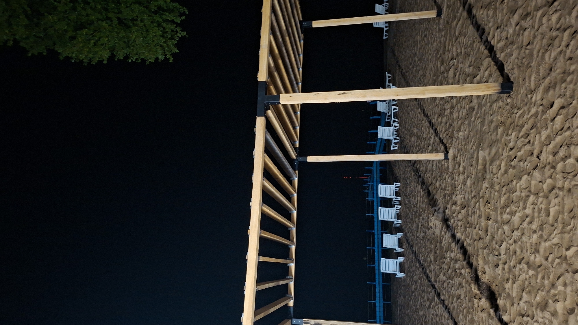 This is a nighttime, medium-angle shot of a sandy beach with a wooden pergola structure in the foreground. The pergola has a slatted roof and wooden posts. Behind the pergola, several white lounge chairs are arranged on the sand. A blue railing runs along the edge of the beach, with a body of water visible beyond it. The sky is dark, and a few trees are visible in the upper right corner. The image appears to be taken in Myadzyel, Belarus.