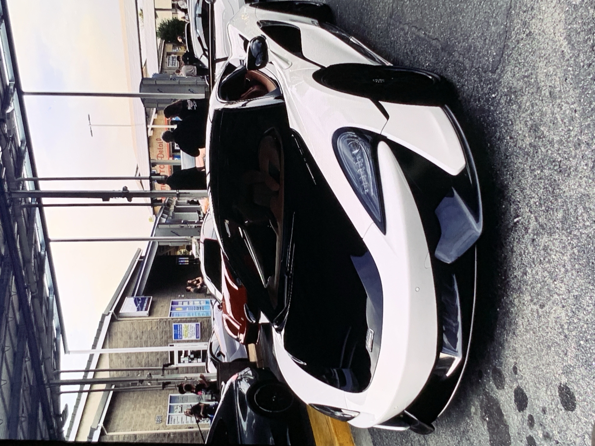 The image captures a white McLaren sports car parked in front of a building with a covered overhang. The car features a black hood and accents, and its doors are open, revealing a tan interior. Several other cars are visible behind the McLaren, including a red sports car and a dark-colored sedan. In the background, people are gathered near the building entrance, some appearing to be interacting. The environment suggests an urban setting, possibly a car detailing shop or a gathering for car enthusiasts. The lighting indicates it might be late afternoon or early evening.