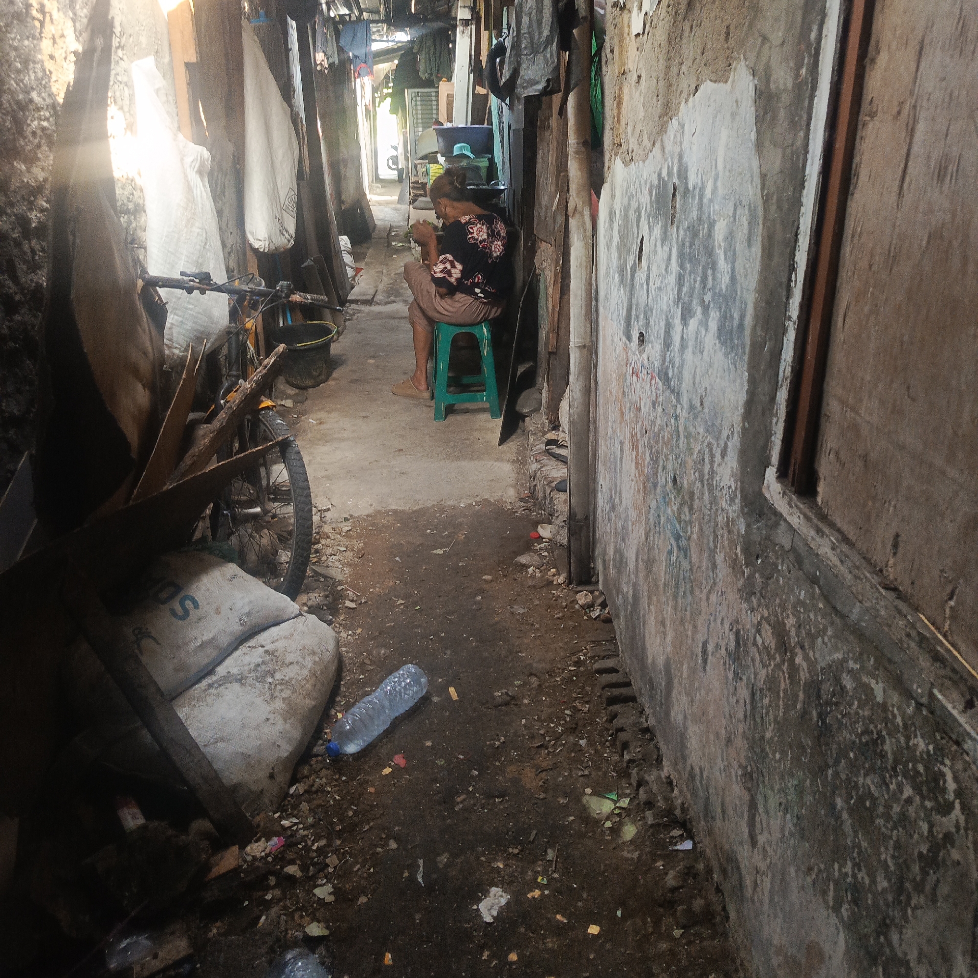 An outdoor alleyway in South Jakarta, Indonesia, is depicted. Sunlight streams in from the left, illuminating a narrow passage between two weathered walls. On the right wall, there are signs of age and disrepair, with peeling paint and stains. The ground is uneven and littered with debris, including plastic bottles and small pieces of trash.
In the middle of the alley, a woman is seated on a green plastic stool, facing away from the camera. She is wearing a dark patterned shirt and light-colored pants. She appears to be engaged in some activity, possibly eating or sorting something.
To the left of the woman, a bicycle is parked, leaning against a pile of objects that include a large sack with "SDS" printed on it and some wooden planks. The alley is also lined with various items, including what appear to be tarps or large fabric sheets hanging from overhead structures, and laundry drying. The overall impression is one of a cluttered, working-class environment.