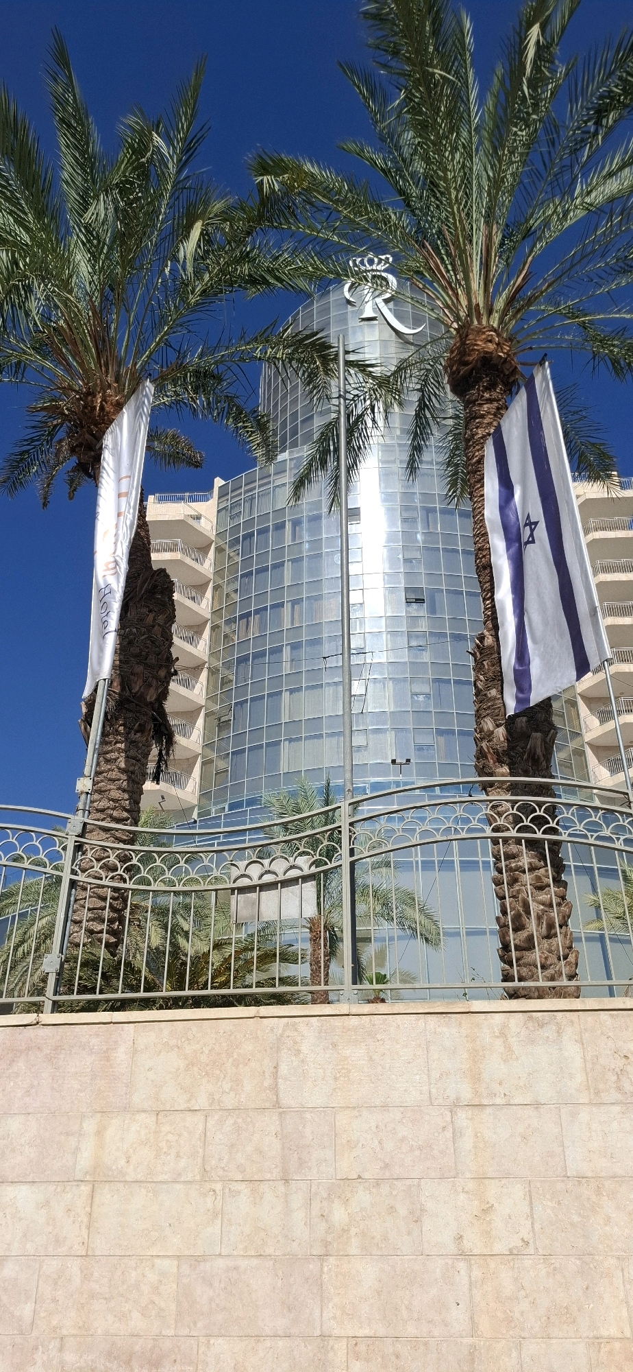 A view of the exterior of a modern, glass-fronted hotel building, likely the Royal Beach Hotel in Eilat, within the Tamar Regional Council, Israel. Two prominent palm trees flank the foreground, with one displaying an Israeli flag and the other a white banner with text. A decorative metal fence separates the foreground from the hotel grounds, where more palm trees are visible. The sky is a clear, bright blue, indicating a sunny day.