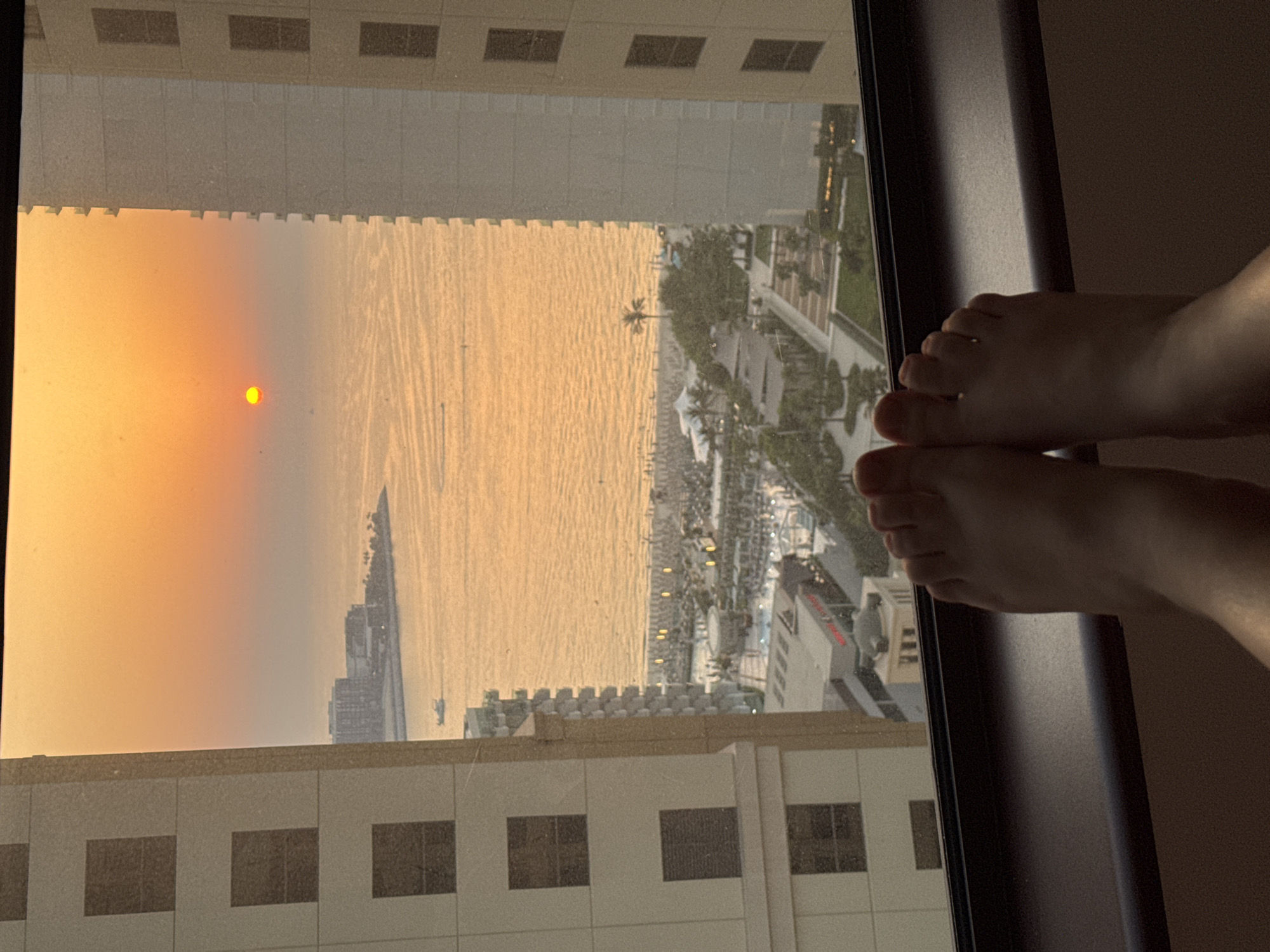 The image captures a view from a high-rise window, overlooking a coastal cityscape during sunset. In the foreground, a person's bare feet are resting on the window ledge, partially obscuring the view. The setting sun casts a warm, orange glow across the sky and illuminates the ocean, creating a shimmering surface with gentle ripples.
The cityscape unfolds below, featuring a beach lined with palm trees and resort-style buildings. Further out, a large cargo ship is visible on the water, silhouetted against the colorful sky. The architecture of the buildings suggests a modern, possibly resort-oriented, urban environment. The overall mood is serene and contemplative, enhanced by the warm hues of the sunset. The time of day is clearly sunset. There is no visible text in the image. Given the architecture and the presence of a large cargo ship and resort areas, the location is consistent with Dubai, United Arab Emirates.