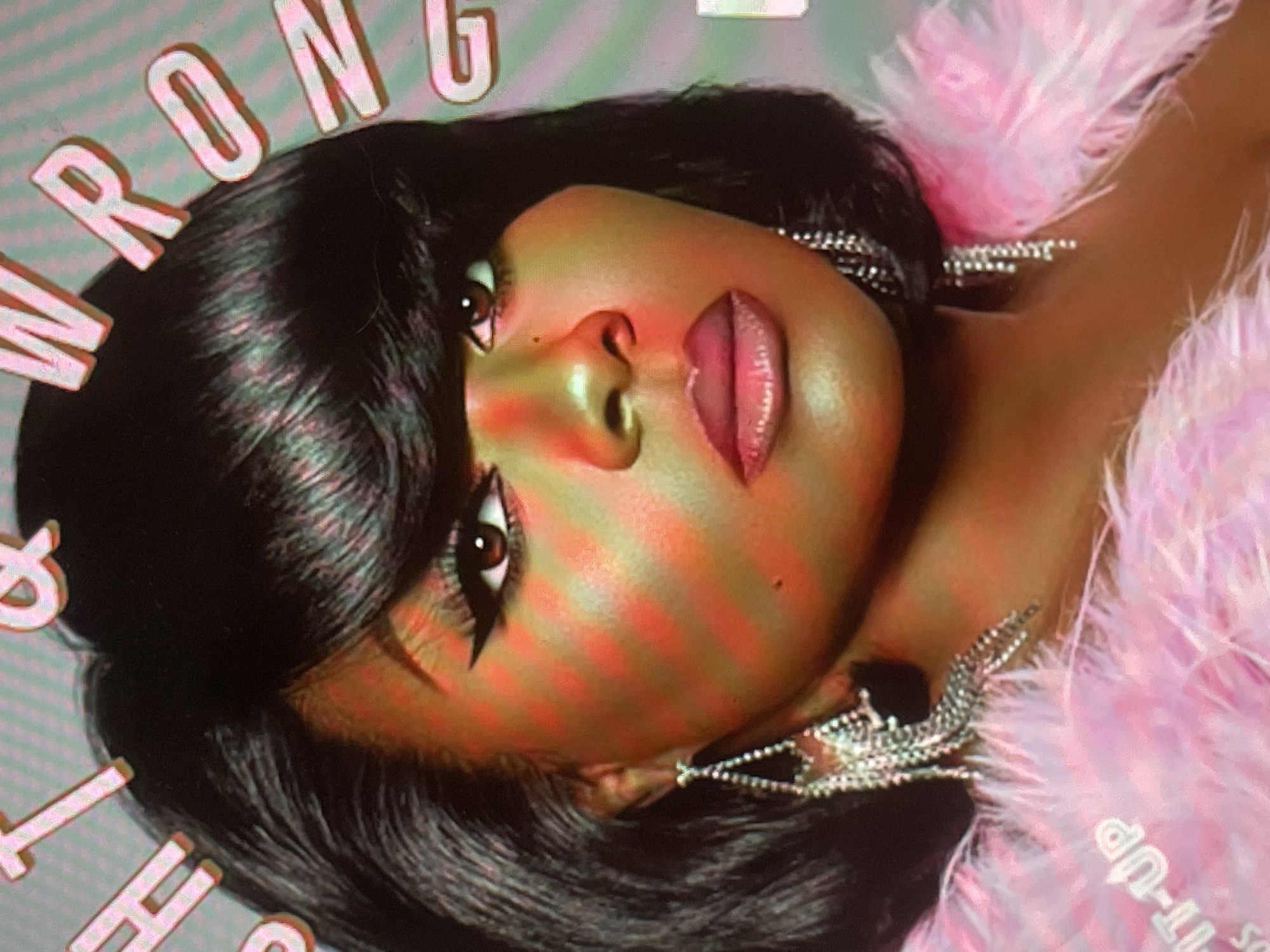 A close-up, tilted portrait features a person, likely a woman, with dark, straight hair and bangs, looking directly at the viewer with an intense, neutral expression. Her face is made up with dramatic winged eyeliner, defined brows, and glossy pink lips. Large, ornate, silver-toned sparkling earrings adorn her ears, and she is wearing or adorned with voluminous pink feathery material around her neck or shoulder. The person's skin shows a warm, golden hue, overlaid with distinct red and orange diagonal light stripes, suggesting specific artificial lighting or reflection from a digital screen. In the background, large, white, three-dimensional letters spelling "W R O N G" are partially visible above her head. Additional partial text, "IT-UP," is visible on the pink feathery material. The scene appears to be an indoor studio setting for a styled photograph, possibly for promotional purposes. No specific time of day or location cues for Sofia, Bulgaria, are visible within the image itself.