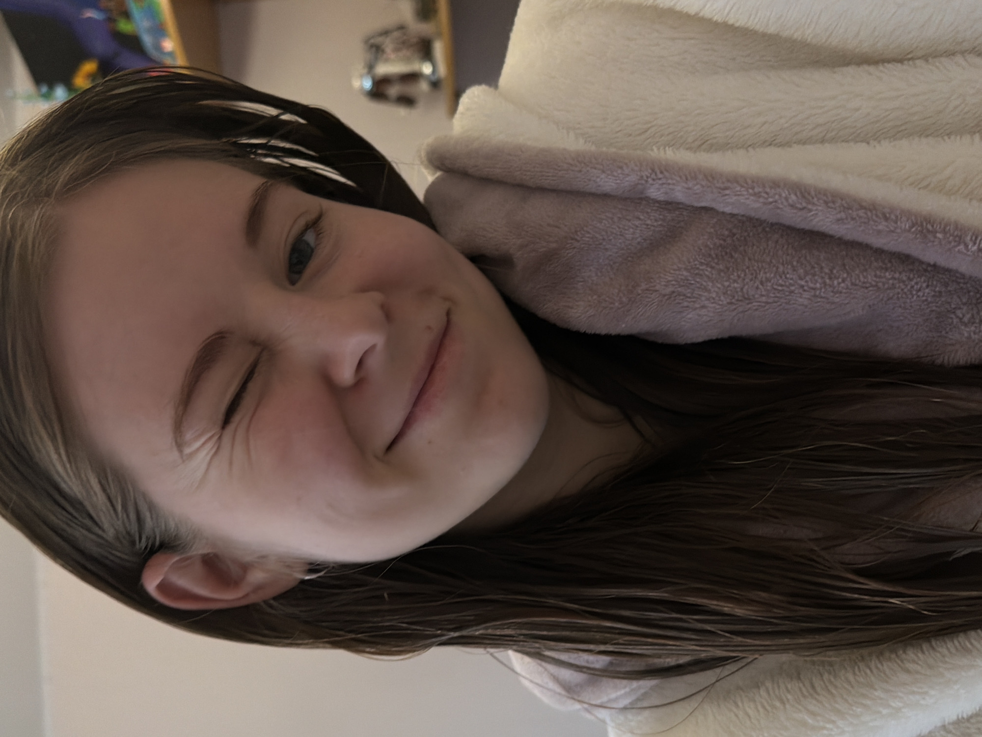 A close-up portrait, originally rotated, features a young person, likely female, with long, dark brown hair that appears sleek or damp. She is winking with one eye while the other remains open, and a subtle, playful smile graces her lips. Her fair skin is visible, with delicate wrinkles apparent around the winking eye. She is partially enveloped in a plush, two-toned garment or blanket, which displays an outer layer of light cream or beige and an inner lining of soft lilac or light purple. The background is intentionally blurred, revealing only indistinct elements of an indoor setting, possibly a room in a home, with some out-of-focus objects. The image conveys a cheerful and casual mood. No visible text is present. The provided context indicates the location is Perm, Russia.