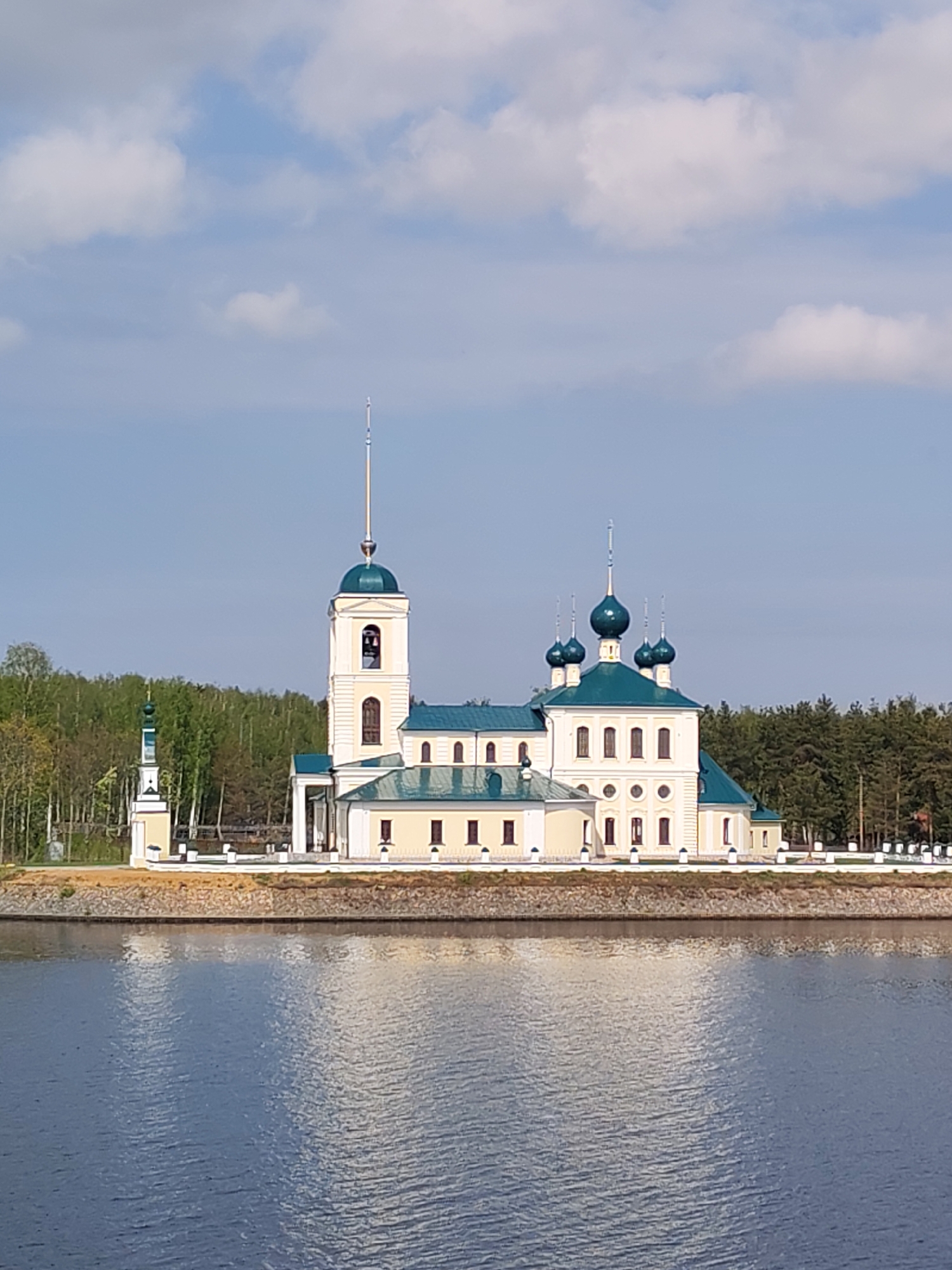 The image shows the Church of Praise of the Mother of God, located in Koprino, Russia. The church is situated on a small landmass surrounded by water, possibly a lake or wide river. The structure is predominantly cream-colored with green roofs and blue domes, typical of Russian Orthodox architecture. The church complex includes a bell tower topped with a spire, and a smaller decorative tower on the left side. A low white fence runs along the perimeter of the property. Behind the church, a dense forest is visible. The sky is blue with scattered white clouds. The water in the foreground reflects the church and the sky.