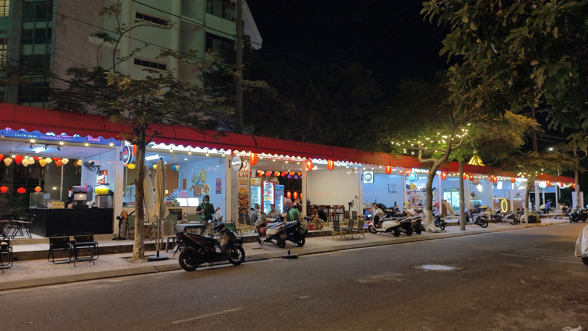 A nighttime exterior view shows a row of food stalls or small restaurants along a city street in Nha Trang, Vietnam. The establishments are set up under a continuous red awning, illuminated by strings of white fairy lights draped over trees and poles, and accented with red and yellow paper lanterns. Each stall features an open-fronted design, allowing visibility into the brightly lit interiors where food is prepared and served. Several people are visible, some seated at small tables outside and others working behind the counters. Scooters are parked along the sidewalk and on the street. The overall atmosphere is lively and suggests a popular dining or takeaway spot.