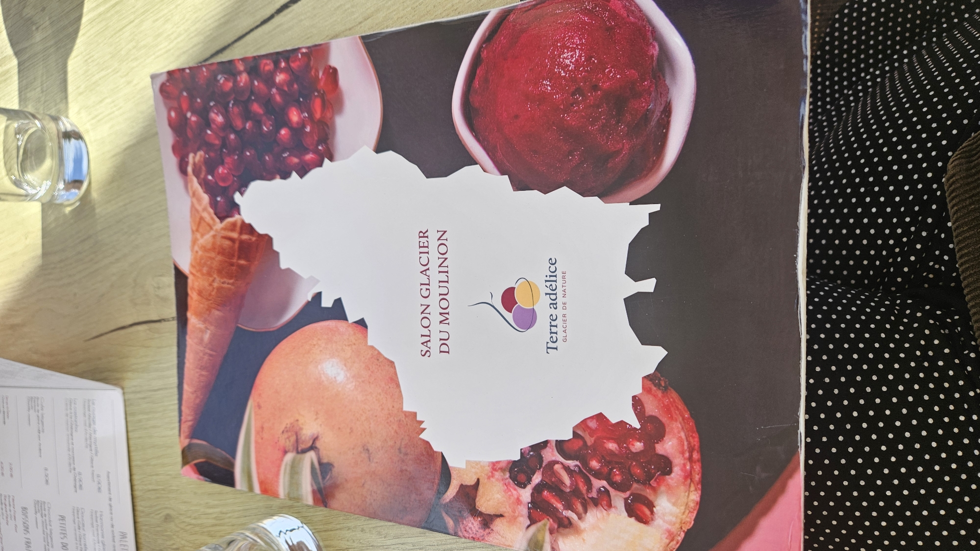 The image shows a close-up, top-down view of an open menu on a wooden table. The menu features images of pomegranates, an ice cream cone, and a scoop of sorbet. The text on the menu reads "SALON GLACIER DU MOULINON" and "Terre adélica GLACIER DE NATURE". A glass of water is visible in the upper right corner of the image. The menu is positioned on a dark fabric with a white polka dot pattern. The scene is set at the Salon Glacier du Moulinon in Saint-Sauveur-de-Montagut, France.