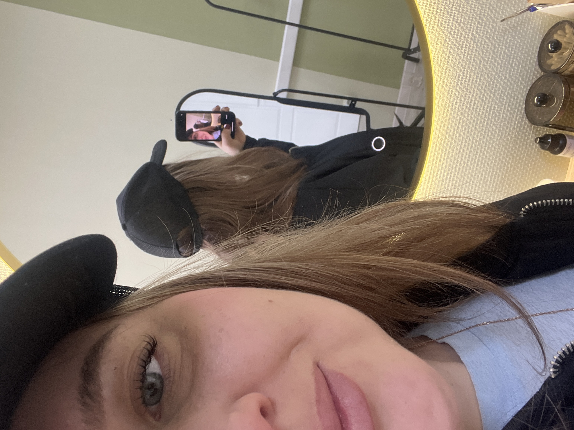 An indoor mirror selfie captures a woman from a tilted, close-up perspective in Chaikovsky, Russia. In the immediate foreground, the right side of the woman's face is visible, showing her eye with long lashes, the side of her nose with a piercing, and slightly parted lips. Her long, light brown hair cascades around her, partially covering a light blue top and a black jacket with a silver zipper. She wears a black baseball cap.
The main subject is looking towards a large, circular mirror with a golden-yellow textured frame, positioned to her right. The reflection in this large mirror shows the woman from behind, wearing the same black cap and jacket, holding a smartphone up to take the selfie. Behind her, a rectangular mirror, possibly a rearview mirror, is also visible. This smaller mirror reflects the screen of her phone, showing the image she is capturing: a recursive view of the large mirror and her reflection.
On a surface next to the main mirror, several small bottles or containers are partially visible, suggesting a beauty or personal care setting. The background visible in the main mirror reflection shows light-colored walls and a hint of a light fixture or overhead structure. The lighting is bright and even, indicating daytime or well-lit indoor conditions. No visible text is legible in the image.