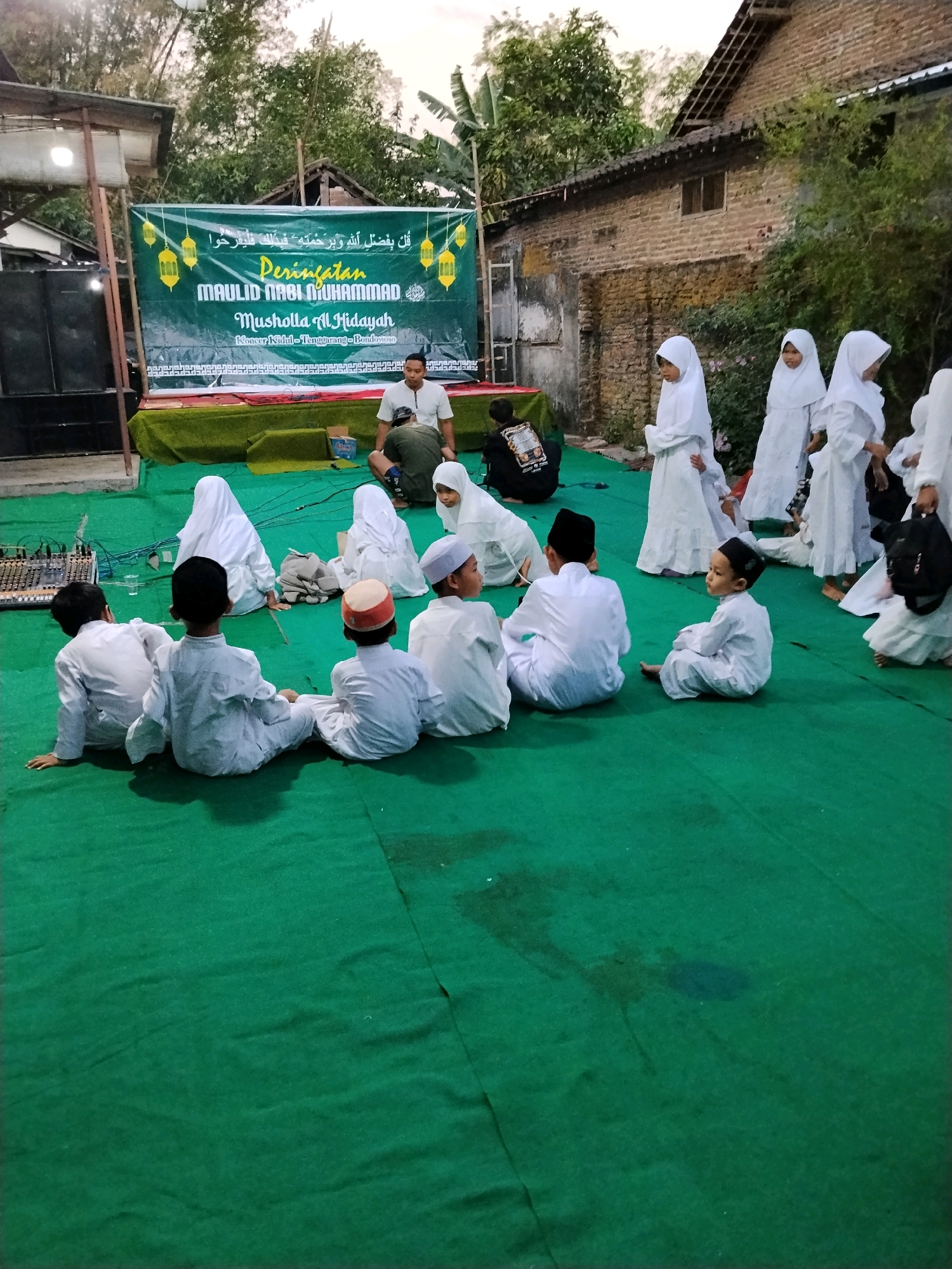 In Kejawan, Indonesia, a group of children, predominantly boys wearing white shirts and black caps or white caps, are seated on a green carpet outdoors, facing a banner. The banner, displayed on a stage with a green backdrop, is primarily green and yellow and features Arabic script and decorative elements. Text on the banner reads "PERINGATAN MAULID NABI MUHAMMAD MUSHOLLA AL-HIDAYAH KECAMATAN GALUR." To the left of the banner, speakers are visible. A few adults, one in a white shirt and another in a dark shirt, are also present, interacting with the children or observing the scene. The environment appears to be an open area with some vegetation and a brick building in the background. The overall scene suggests an outdoor religious gathering or event, possibly a celebration related to the Prophet Muhammad's birthday.