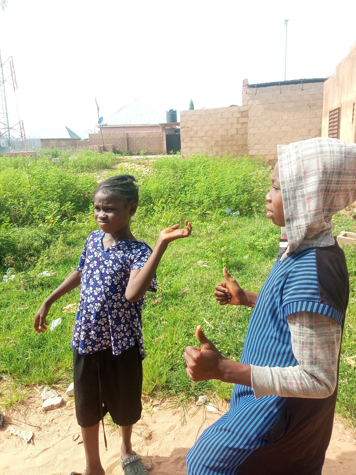 In Tempure, Nigeria, two young people are standing in a dusty, outdoor area. The person on the left, a child with dark braided hair, wears a blue shirt with a white floral pattern and black shorts. They are looking to their left with a slightly questioning expression and their right hand is raised, palm open. The person on the right, partially obscured by the frame, is wearing a blue and black striped garment with a grey and white checkered head covering. Their right arm is bent, and their thumb is raised in a "thumbs up" gesture. In the background, there is lush green vegetation, a brick building, and a tall communication tower. The ground is sandy and uneven, with some scattered debris.