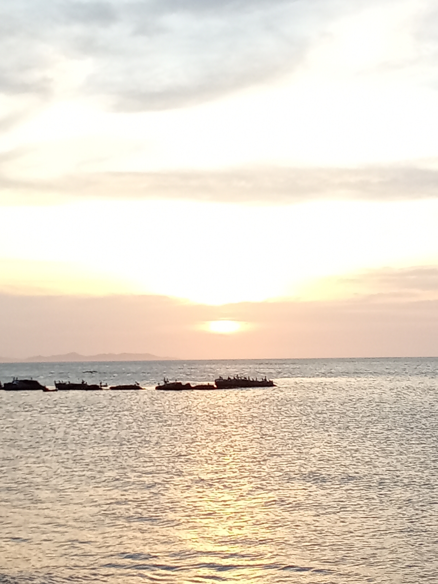 The image shows a sunset over the ocean in Juan Griego, Venezuela. The sky is light with clouds. The sun is setting on the horizon. Several connected boats line the horizon, with objects and people on them. The water reflects the sunlight, creating a shimmering effect.