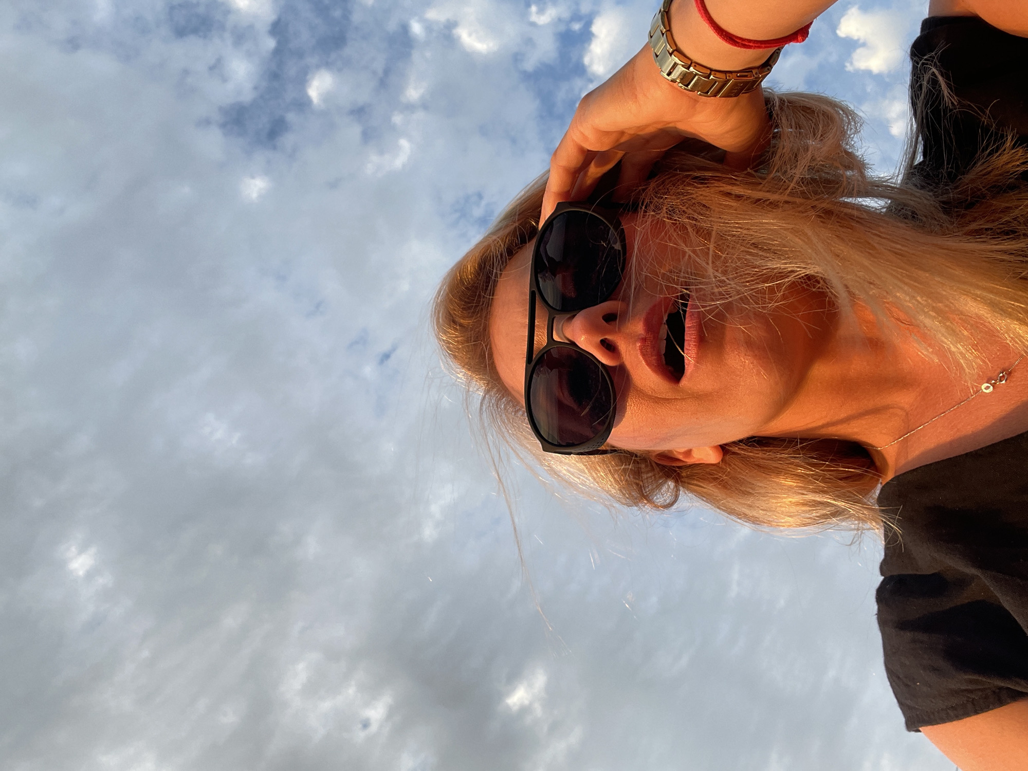 A low-angle, close-up shot captures a woman from the chest up against a cloudy sky. She has blonde hair and is wearing sunglasses and a black top. Her right hand is raised, adjusting her sunglasses. A gold watch is visible on her right wrist, along with a red string bracelet. Her lips are slightly parted, and her mouth is open. The lighting suggests it is either sunrise or sunset, casting a warm glow on her face. The background consists of a mottled sky with scattered clouds, some appearing light and wispy, while others are darker and more defined. There are no discernible city landmarks or specific environmental features that would indicate a location in Unknown, Unknown.