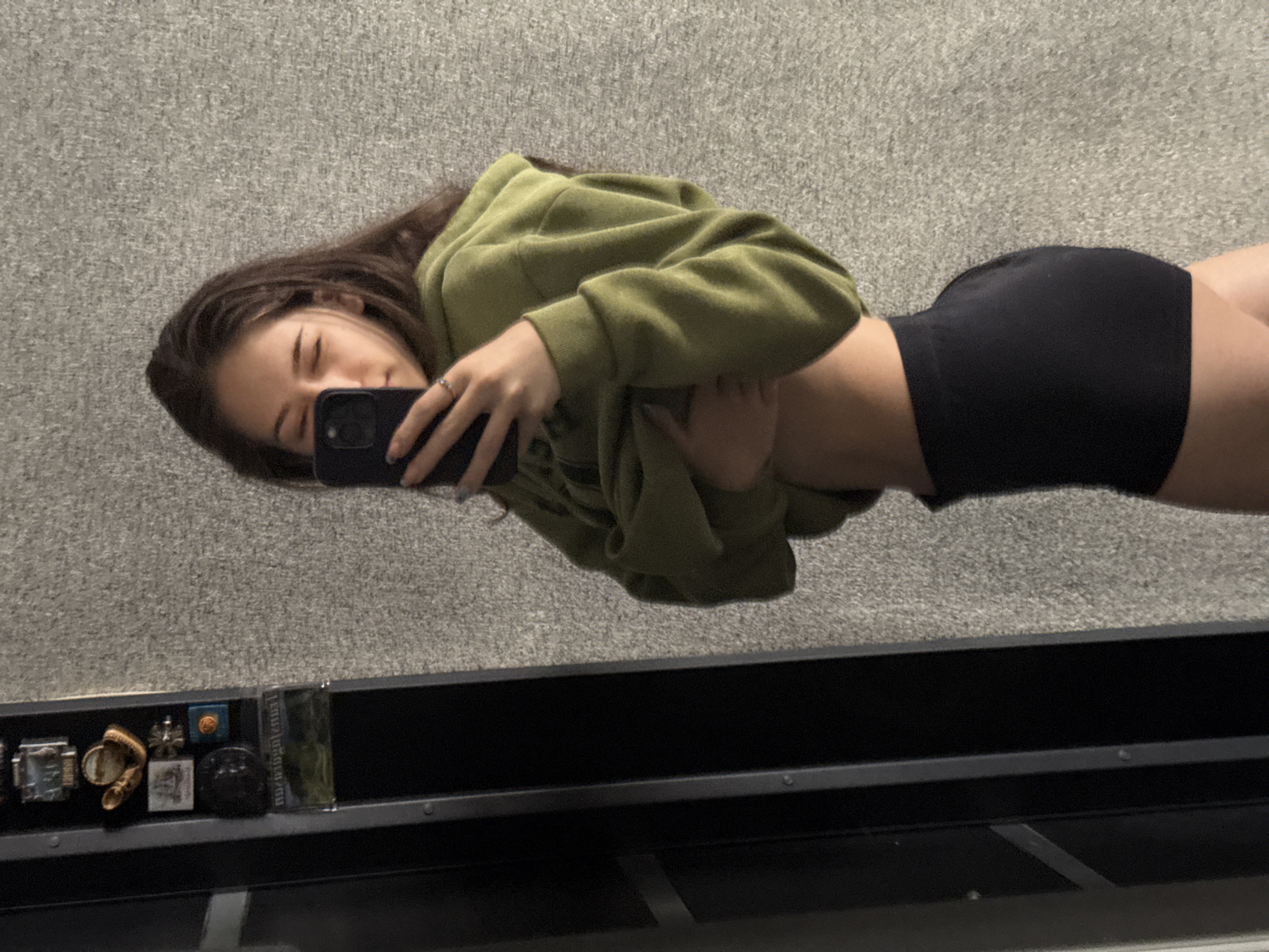 A young person is captured taking a mirror selfie in an indoor setting in Sofia, Bulgaria. The image depicts the subject lying down or leaning back against a light grey, textured surface, resembling carpet or a soft wall.
The subject has long dark hair and is wearing an olive green hooded sweatshirt (hoodie) and black shorts or underwear. They are holding a dark-colored smartphone with both hands, positioned in front of their midsection, aiming the camera at themselves. Their eyes are mostly closed, suggesting a relaxed, dreamy, or possibly playful expression. A ring is visible on the ring finger of their left hand. The hoodie is partially pushed up, revealing a section of their midriff.
In the reflection below the person, a black shelf or ledge is visible, neatly holding several small objects. From left to right, these include a metallic rectangular item, a small light-colored braided or woven object, a silver cross-like emblem, a small blue box with an orange square on top, a black circular item, and a transparent rectangular object (possibly a cassette tape or small book) with faint, undecipherable text on its side. The background behind these items is the same grey textured surface. The lighting appears to be ambient indoor light.