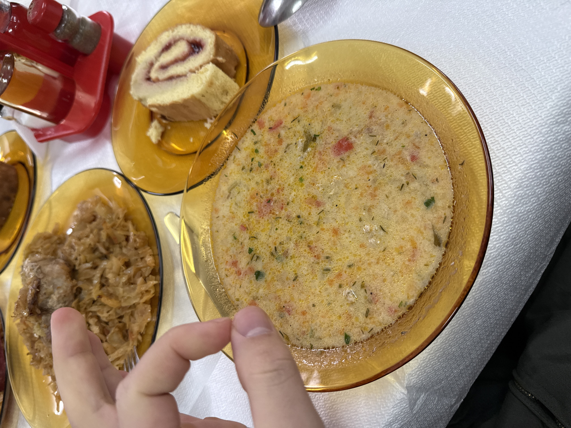 On a white tablecloth, a meal is laid out consisting of a bowl of creamy soup in an amber glass bowl, a plate of sliced swiss roll cake, and a plate of stewed cabbage with meat. A hand reaches towards the cabbage. A red condiment holder with what appear to be salt and pepper shakers sits to the side. The location is likely Sofia, Bulgaria, based on the types of food shown.