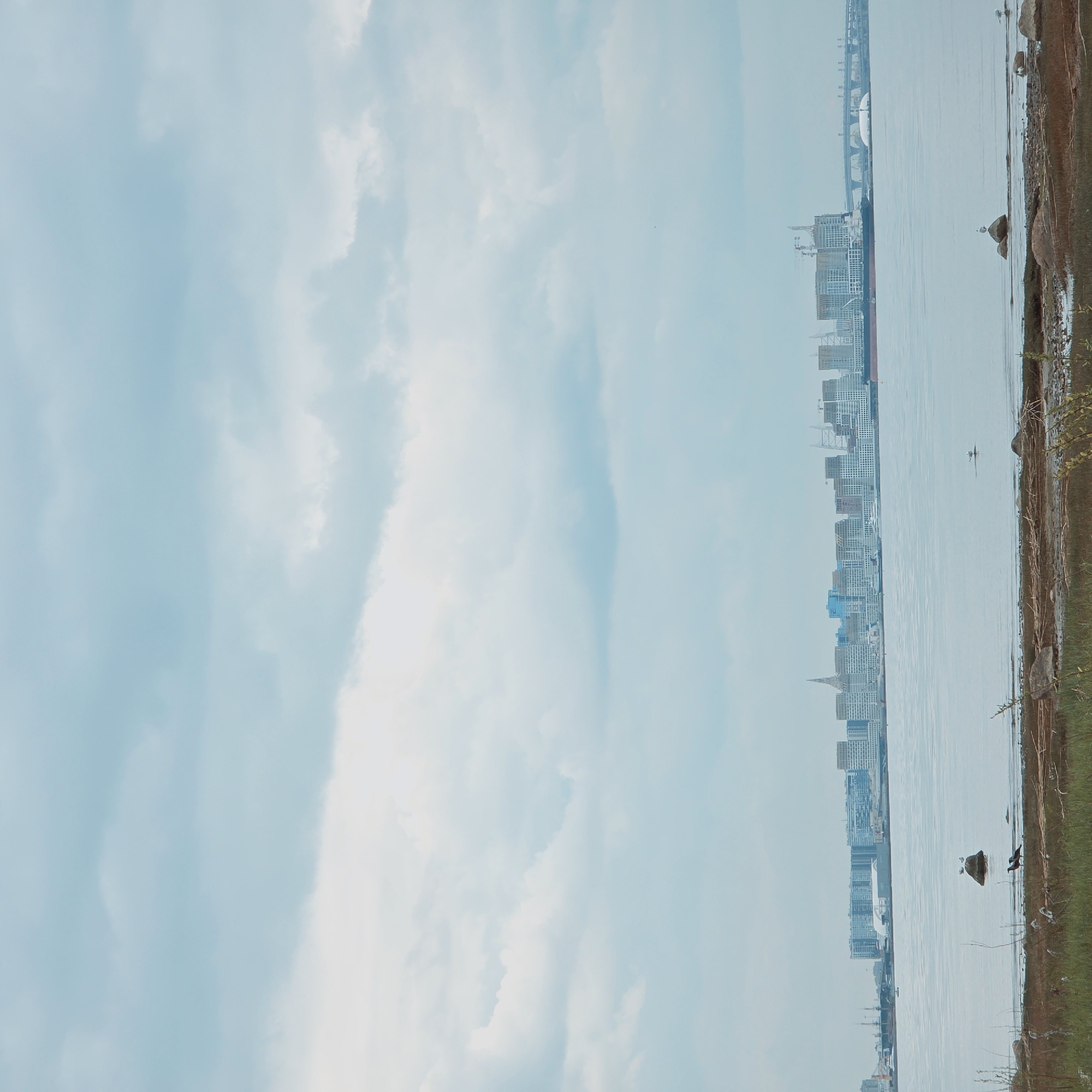 A vertical shot shows a cityscape in the background, with the foreground divided between the sky and water, and a small strip of land. The sky, filling the left side, is light blue with scattered white clouds. The water stretches across the middle of the image, with a smooth surface. The right side is taken up by a strip of land, covered in grass and rocks. Along the horizon, the cityscape is visible, with tall buildings and structures.