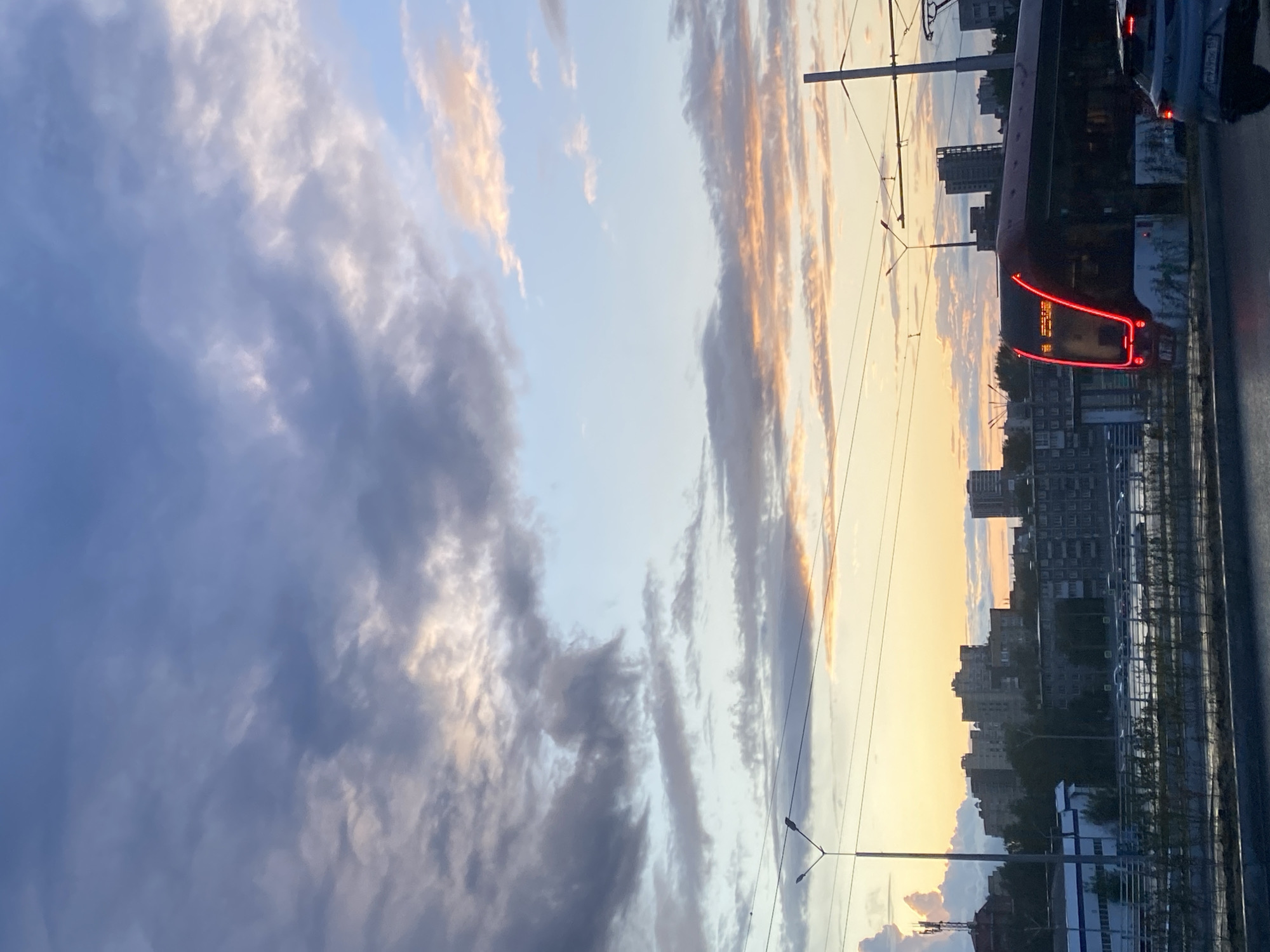 The image depicts a city scene in Perm, Russia, during sunset. A modern tram with illuminated red headlights is prominently featured on the right side, moving from left to right. The tram has an electronic display on its front showing text, likely route information. In the background, a cityscape with various buildings, including apartment blocks, is visible. The sky is filled with dramatic clouds, showcasing hues of orange, pink, and blue, indicative of a sunset. Power lines and streetlights are also present, suggesting an urban environment. The foreground shows a road and some vegetation. No individuals are clearly visible in the image.