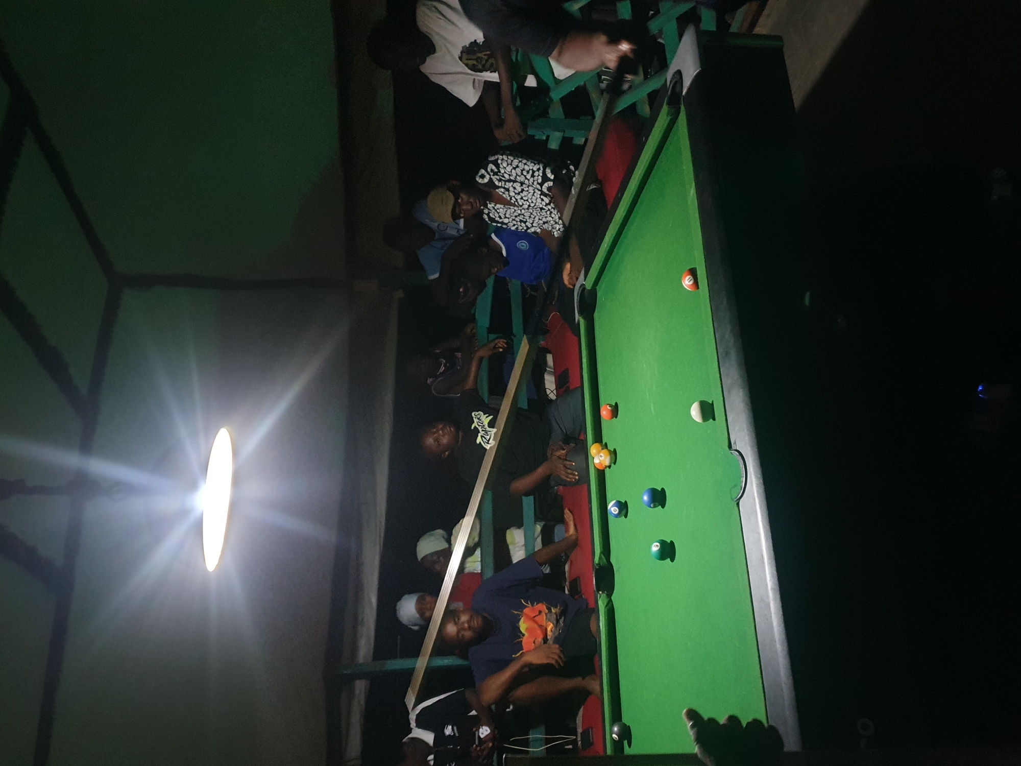 The image captures a dimly lit indoor scene, likely a recreation area or a bar, situated in Lokoja, Nigeria. A group of individuals are gathered around a green pool table, some actively playing and others observing. Overhead, a bright light fixture illuminates the space, casting dramatic shadows. The atmosphere appears casual and social, with people dressed in everyday clothing.
In the foreground, a pool table with several balls is visible. The balls are of various colors, including white, black, blue, orange, and yellow, and some are numbered. The table has a green felt surface and dark green cushioned sides. Two individuals are positioned close to the table, one in a kneeling posture and the other with his legs extended, seemingly engaged in the game.
In the midground, several other people are seen watching the game or conversing. They are seated or standing behind the pool table. Among them, some are wearing head coverings. The background is mostly obscured by darkness, but suggests a rustic or outdoor structure, possibly with wooden beams and supports visible overhead. The overall impression is of a community gathering for leisure and entertainment.