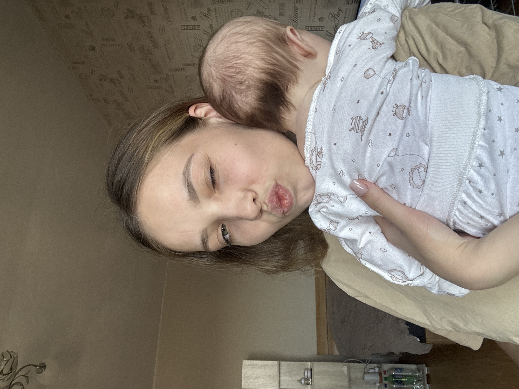 A person with shoulder-length brown hair is holding a baby. The baby is wearing a white outfit with animal prints. The person is making a kissing face toward the camera. The scene is indoors. Underneath the person is furniture. A wall is partially visible to the left and above, with decorative wallpaper. Near the bottom of the image, there is a section of a shelf, and bottles containing green liquids can be seen. The location may be in Chaikovsky, Russia, given the instructions provided.
