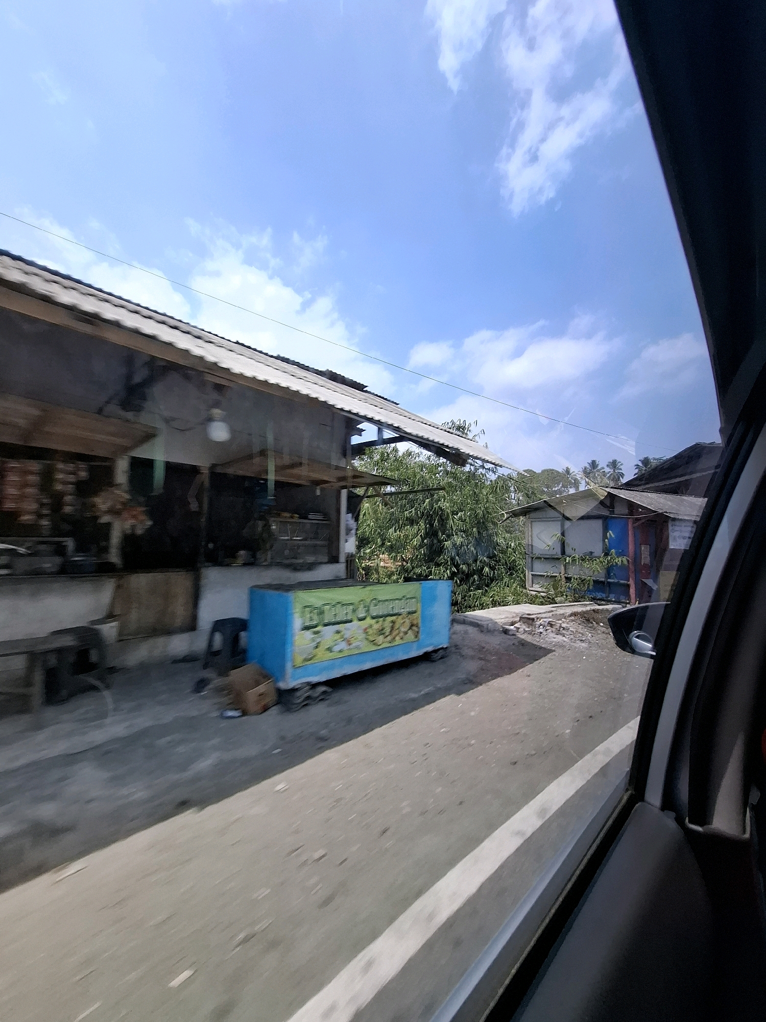 The image captures a roadside scene in Cijangkar, Indonesia, on a sunny day with a clear blue sky and scattered clouds. On the left, a long, low building with a corrugated metal roof houses a shop or market. Shelves inside display various goods, and an open display area suggests food or household items. In front of the building, a blue refrigerated display case, likely for drinks or food, is situated on the pavement. The pavement itself is a mix of gravel and what appears to be asphalt. To the right of the shop, a narrow road or path curves away. In the background, more structures with similar roofing are visible, along with dense green foliage and palm trees, indicating a tropical environment. A dark-colored car interior is visible along the right edge of the frame, suggesting the photo was taken from inside a vehicle looking out. The scene appears to be a typical street-level view of a commercial area in a rural or semi-urban Indonesian setting.