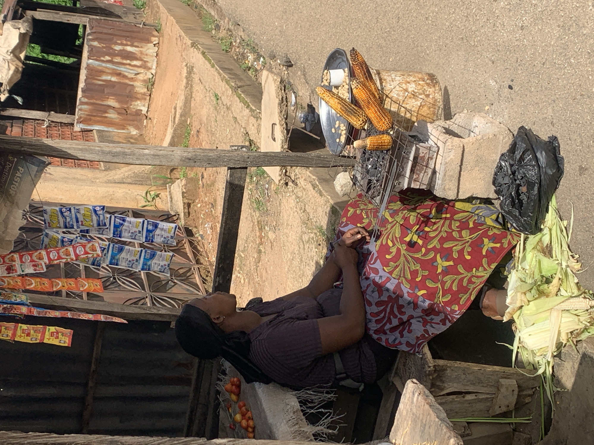 In Jos, Nigeria, a woman sits by a roadside stall, tending to corn roasting on a grill. She wears a dark purple shirt and a vibrant red and yellow patterned skirt. Her head is covered by a dark headscarf. In front of her, corn cobs are arranged on a metal grill atop a brick structure, with glowing embers visible underneath. To the right of the grill, a metal tray holds roasted corn and a small white cup filled with peanuts. A pile of corn husks and stalks lies on the ground to the left, next to a black trash bag. Behind the woman, a stall is decorated with colorful packets of goods, and a corrugated metal structure forms part of the stall's wall. The scene is set on a dirt and concrete ground next to a paved road.