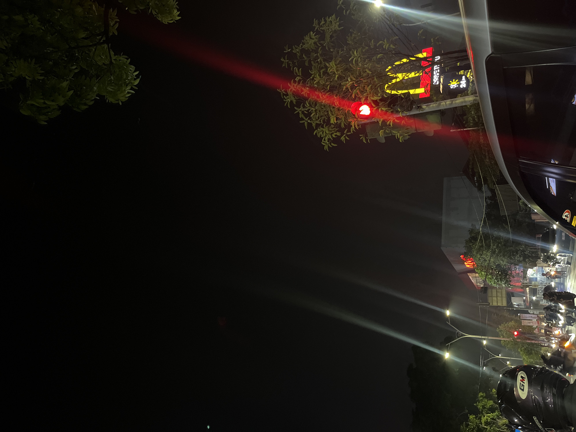 The image depicts a nighttime scene in West Jakarta, Indonesia, likely on a street. In the foreground, the back of a person wearing a black motorcycle helmet with a "GM" logo is visible. Beyond the helmet, a traffic light glows red. To the right, a McDonald's sign with its iconic golden arches is illuminated in yellow against a red background. Adjacent to the McDonald's sign, a smaller sign displays a sun icon and "24 Jam" (24 Hours), indicating the establishment's operating hours. The street is populated with motorcycles and a car, suggesting traffic. Buildings and trees are visible in the background, illuminated by streetlights and the glow from businesses. The overall atmosphere is that of a busy urban street at night.