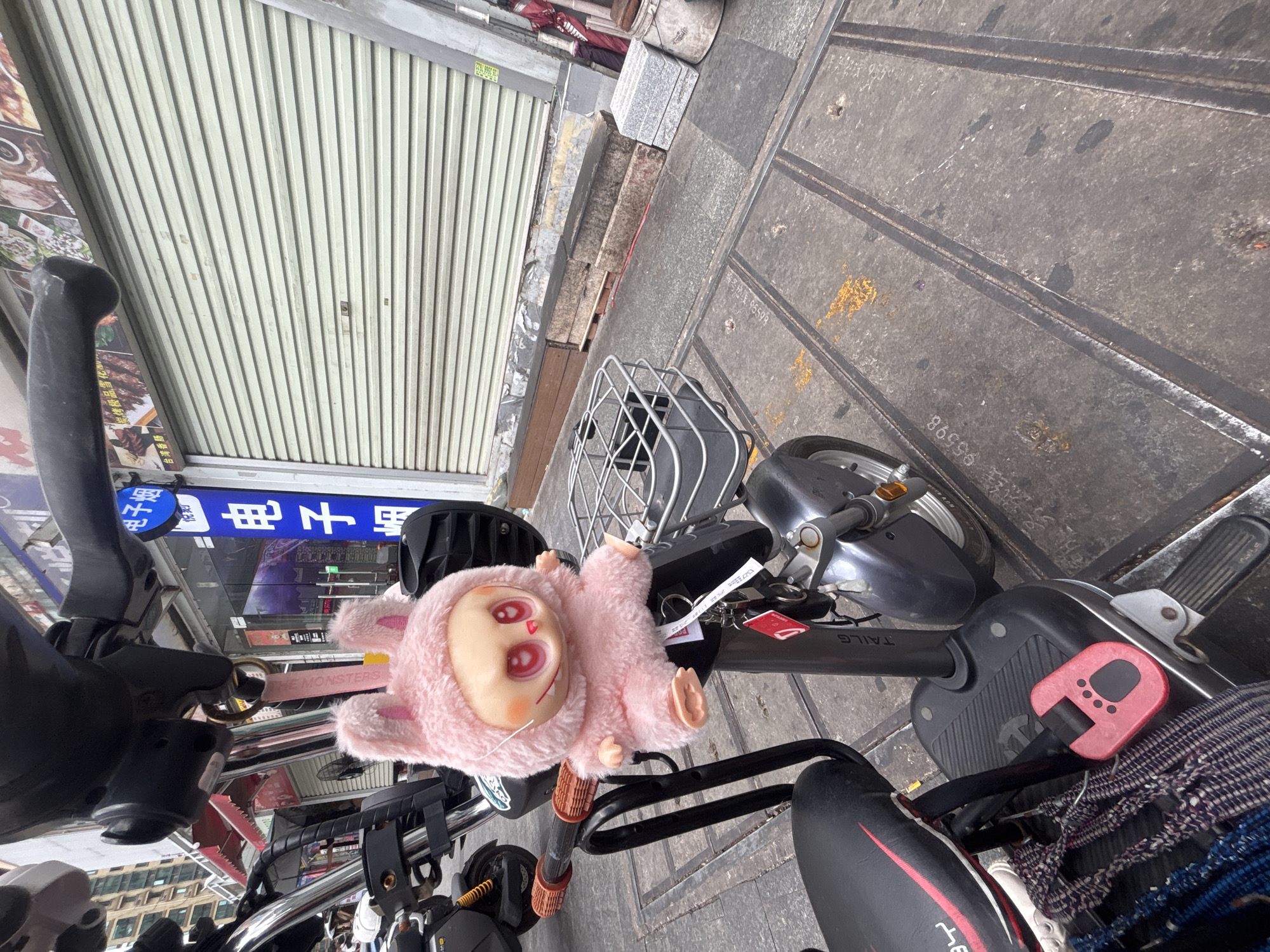 A pink plush toy with rabbit ears and large, red eyes is perched on the handlebars of a parked electric scooter on a city street in Nanhai District, China. The scooter is gray with a black seat and a metal basket attached to the front. In the background, a closed metal roll-up door of a shop is visible, with a sign above it displaying Chinese characters. The street is paved with concrete and has metal tracks embedded in it. Other objects in the scene include a pink pet stroller, bags, and a bucket. The day appears overcast.