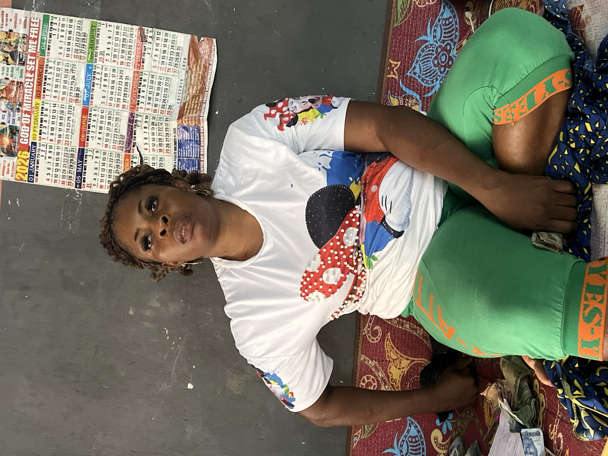 A woman with braided hair, wearing a white t-shirt with a cartoon character print and green pants, sits on a mat. Behind her, a calendar for the year 2026 is pinned to a wall. The calendar has a religious theme, with phrases like "GOD OF MIRACLE SET ME FREE." Scattered around the woman are what appear to be banknotes. The scene suggests a domestic or informal setting, possibly in Nkpor, Nigeria, given the presence of currency and the calendar's context.