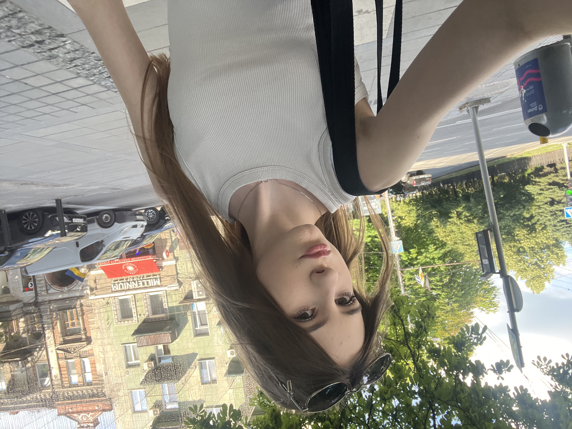 A young woman is captured in a selfie in front of the Millennium Chocolate Store on Karl Marks Avenue (now Velyka Dymerka Street) in Dnipro, Ukraine. She is wearing a white tank top and a black strap from a bag. She has sunglasses perched on her head and her long brown hair is falling over her shoulders. The store's red awning and the yellow and blue Ukrainian flag are visible in the background, along with a white van and other vehicles parked on the street. The scene is set on a sunny day, with trees and buildings lining the avenue.