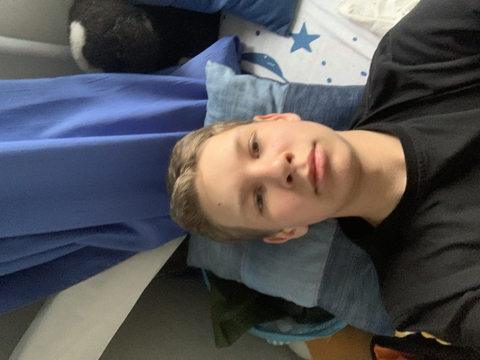A person is lying on a bed. The bed is covered with blue and white bedding, including a pillow with a blue star design. The person is wearing a black t-shirt. A black and white stuffed animal is visible near the top of the frame. The location is Chaikovsky, Russia.