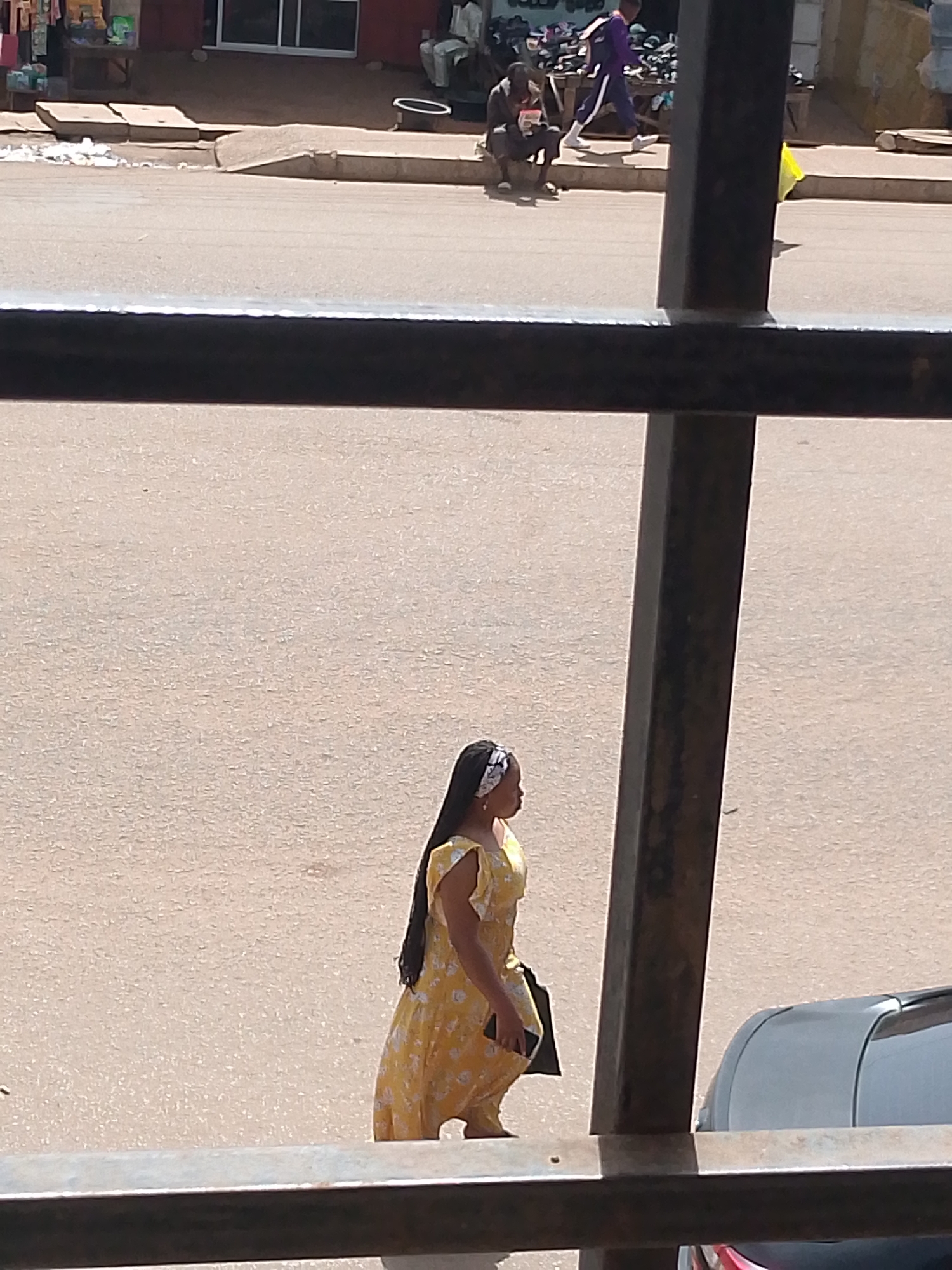 In Bukuru, Nigeria, a woman in a yellow floral dress walks across a paved street. She has a white headband in her hair. In the background, several people are visible, including a man sitting on a curb and another walking in a purple tracksuit. A yellow plastic bag is on the ground. The scene appears to be near shops or market stalls.
