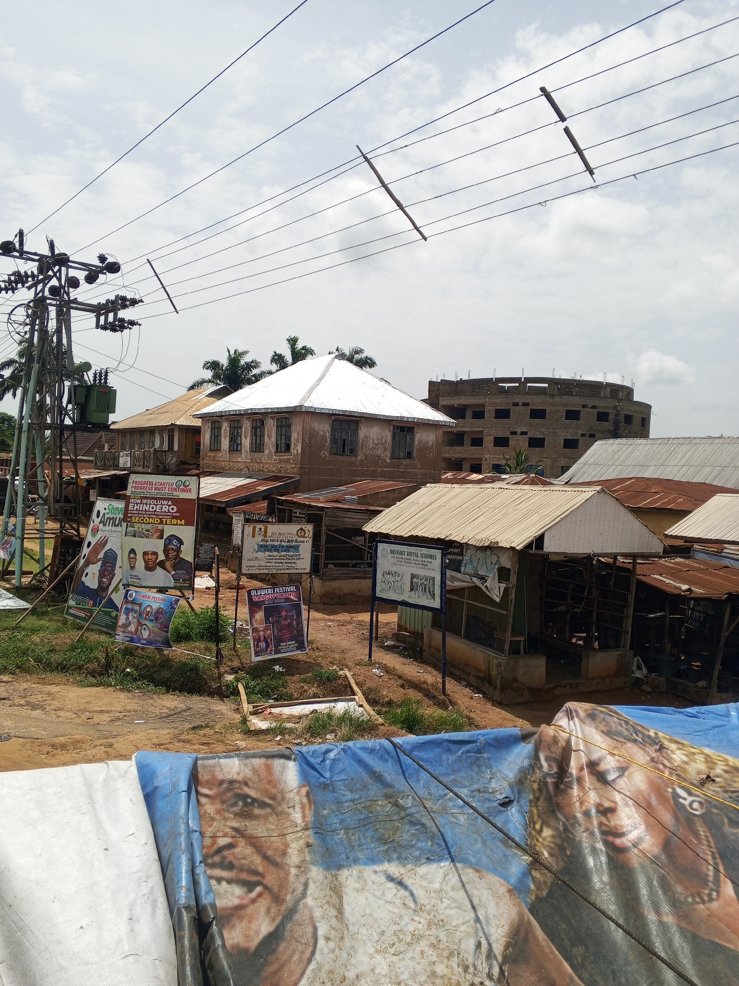 The image depicts a street scene in an urban area of Unknown, Unknown. In the foreground, a blue and white tarp with images of people is stretched out. Behind the tarp are several small, rusty-roofed structures. Further back, there are two older buildings, one with a white roof. A larger, multi-story building under construction is visible in the background. Political posters and banners are displayed along the street, advertising various individuals and events such as "OLUWERI FESTIVAL SANGOFUNSHO DAY 2026". A utility pole with power lines is visible on the left side of the frame. The sky is overcast.
