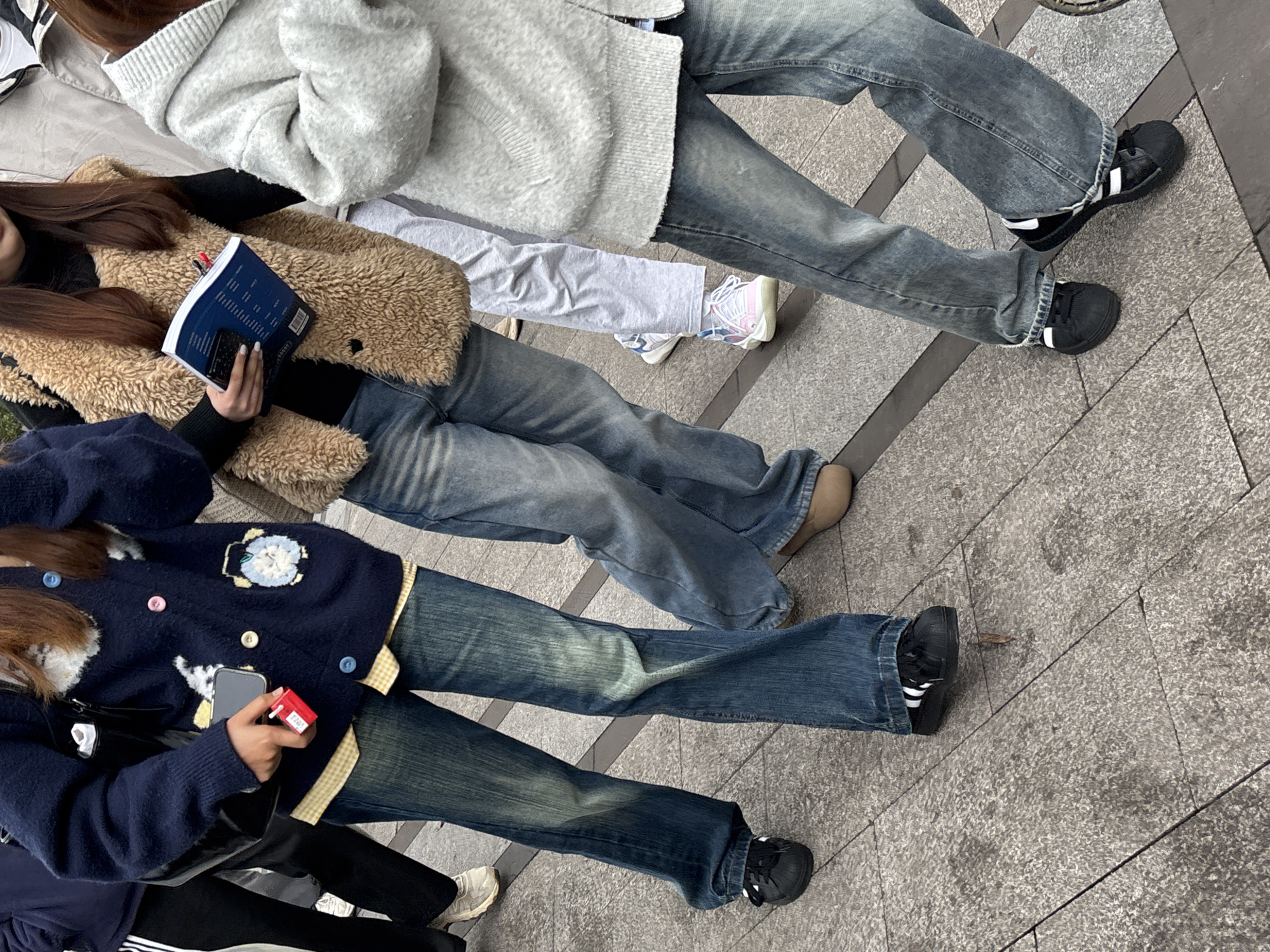 The image shows the lower bodies of three individuals standing on a paved surface with diagonal lines, possibly a public walkway in an urban setting. From top to bottom, the first individual is wearing blue jeans and black shoes with white stripes, a light grey sweater. The second individual is wearing blue jeans, brown shoes and is holding a book. The third individual is wearing blue jeans, black shoes with white stripes and a dark blue sweater. The location is identified as Jiangjin, China.