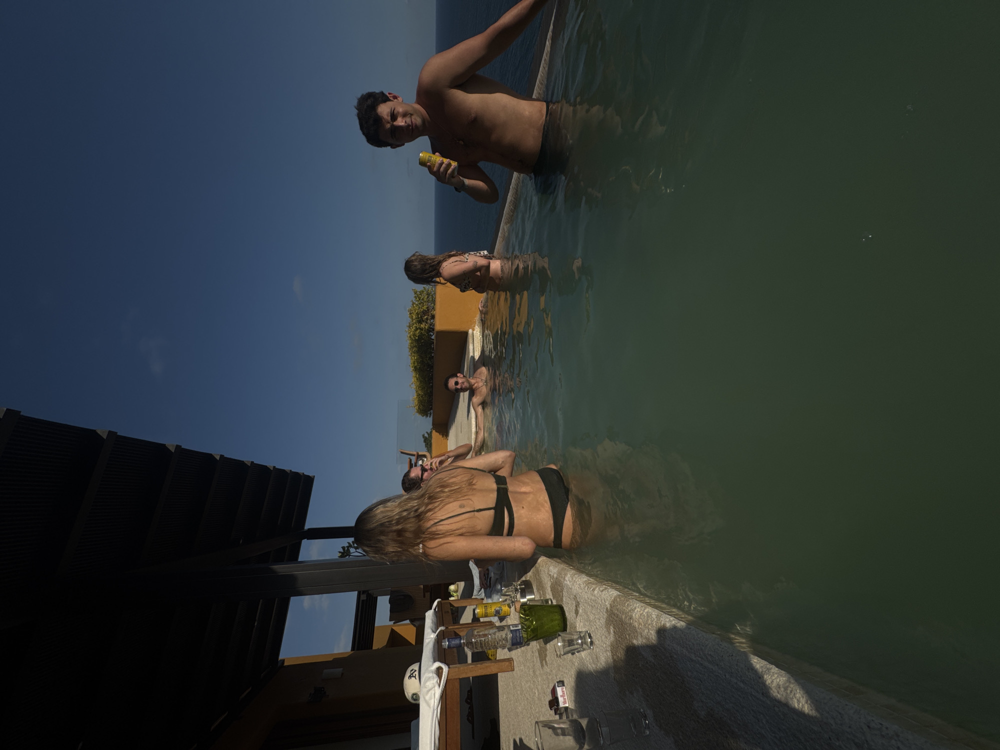 An outdoor scene in Puerto Marqués, Mexico, depicts several people relaxing in an infinity pool under a clear blue sky. The image captures a bright, sunny day, with strong light and shadows.
In the foreground, a woman with long, light brown hair, wearing a dark bikini, faces away from the camera, leaning against the pool's edge. Her lower body is submerged in the greenish water. On the poolside ledge beside her, various items are placed: a clear bottle (likely spirits), a green pitcher, multiple glasses, a yellow beverage can, a white towel, and a red and white cigarette pack with "Marlboro" visible.
In the mid-ground, a shirtless man with dark hair holds a yellow can, looking towards the viewer with a slight smile. He is submerged from the waist down. Behind him, another person in a bikini is seen from the back. Further back, a man wearing sunglasses is partially submerged in the pool.
The infinity pool offers a clear view of the ocean stretching to the horizon. A dark, slatted roof structure on the left provides shade over part of the pool area, hinting at a resort or lounge setting. The clear sky and bright daylight suggest a leisurely daytime activity. The overall scene conveys relaxation and enjoyment.
